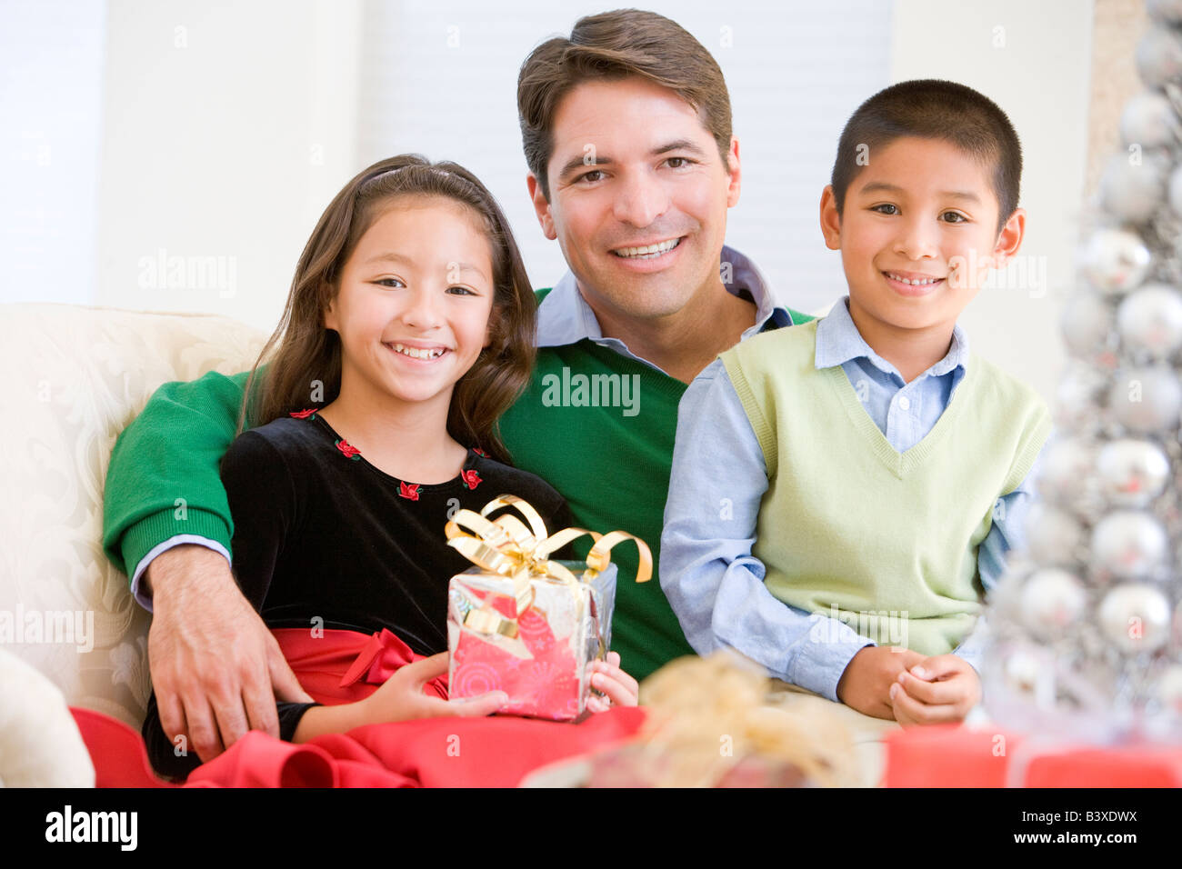 Father And His Son And Daughter Sitting On Sofa, Holding A Christmas Present Stock Photo