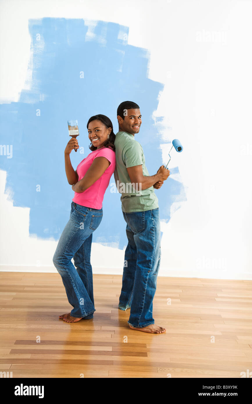 African American couple next to half painted wall as they stand back to back with paint utensils Stock Photo