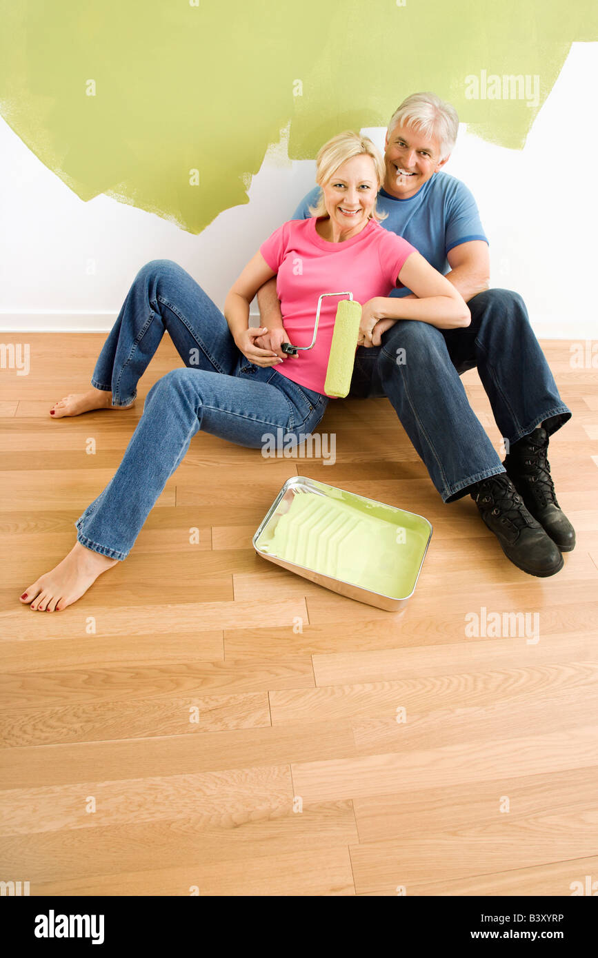 Portrait of happy adult couple sitting in front of half painted wall with paint supplies snuggling Stock Photo