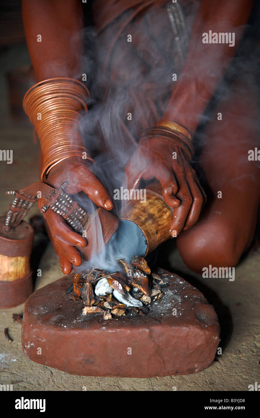 Namibia, Kaokoveld, Himba Village. A Himba woman lights a small fire made of woodshavings and scented butterfat as part of her personal hygiene routine. Once the shavings are smoking she will cover herself with a sheepskin cloak and stand over the fire, while the smoke fumigates lice and leaves her sweet smelling. Stock Photo