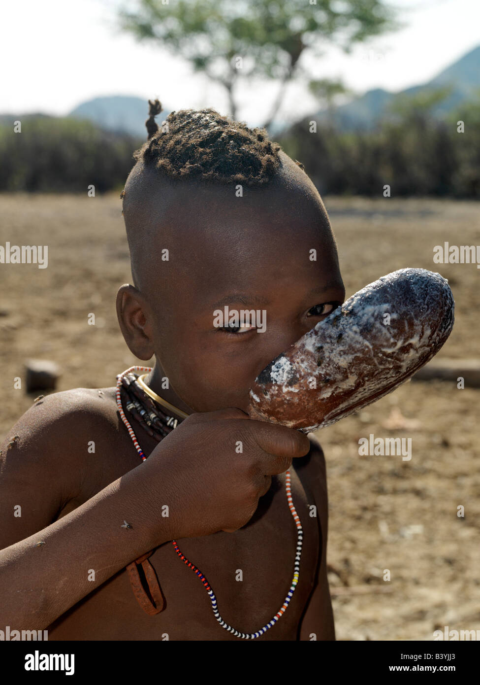 Namibia, Kaokoland. A Himba boy drinks milk from a wooden spoon. His hair is already being styled in a long plait, known as ondatu, which he will keep until he marries.The Himba are Herero-speaking Bantu nomads who live in the harsh, dry but starkly beautiful landscape of remote northwest Namibia. Stock Photo