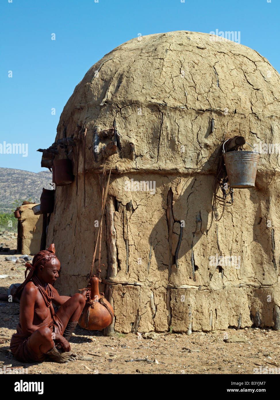 Namibia, Kaokoland. An old Himba woman gently shakes a large milk gourd to make butter outside her home. Her body gleams from a Stock Photo