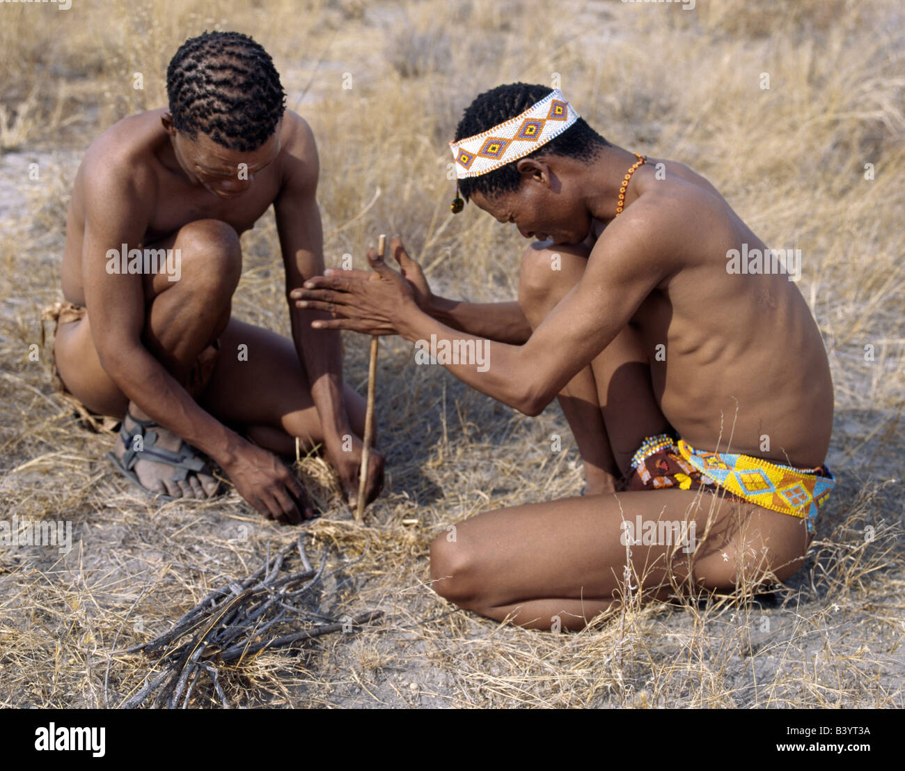 Namibia, Eastern Bushmanland, Tsumkwe. Two !Kung hunter-gatherers light a fire using firesticks. The !Kung people are a part of the San, often referred to as Bushmen. They differ in appearance from the rest of black Africa having yellowish skin and being lightly boned, lean and muscular. They speak with four distinct click consonants.The !Kung live in the harsh environment of a vast expanse of flat sand and bush scrub country straddling the Namibia-Botswana border. Until recently, their way of life had remained unchanged for thousands of years. Few now live solely by hunting and gathering. Stock Photo
