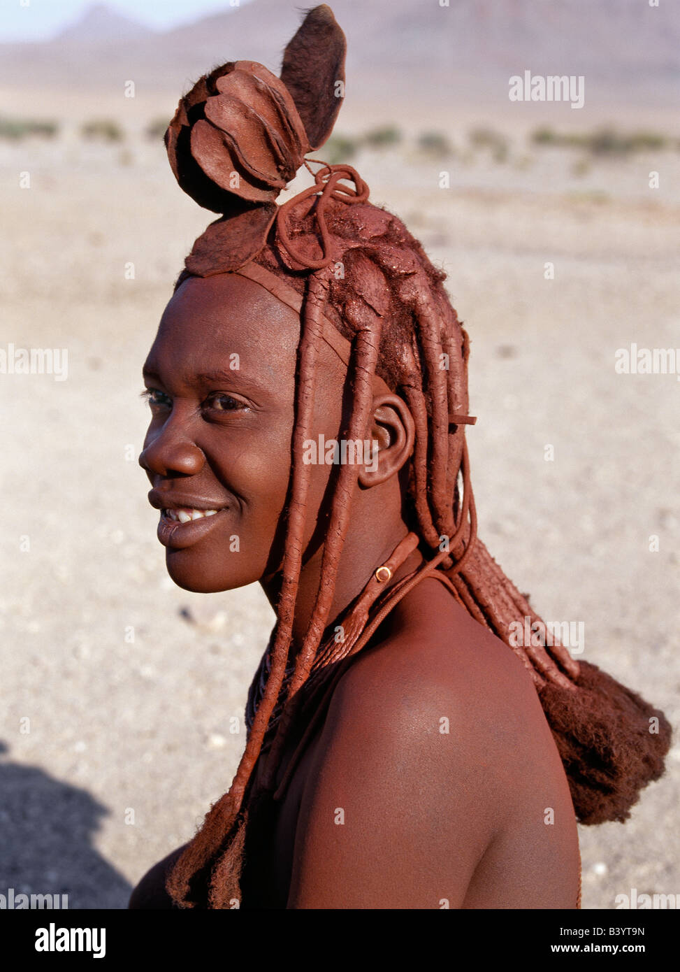 Namibia, Kaokoland, Purros. A Himba woman in traditional attire. Her body gleams from a mixture of red ochre, butterfat and herbs. Her long hair is styled in the traditional Himba way and is crowned with a headdress made of lambskin, called erembe.The Himba are Herero-speaking Bantu nomads who live in the harsh, dry but starkly beautiful landscape of remote northwest Namibia. Stock Photo
