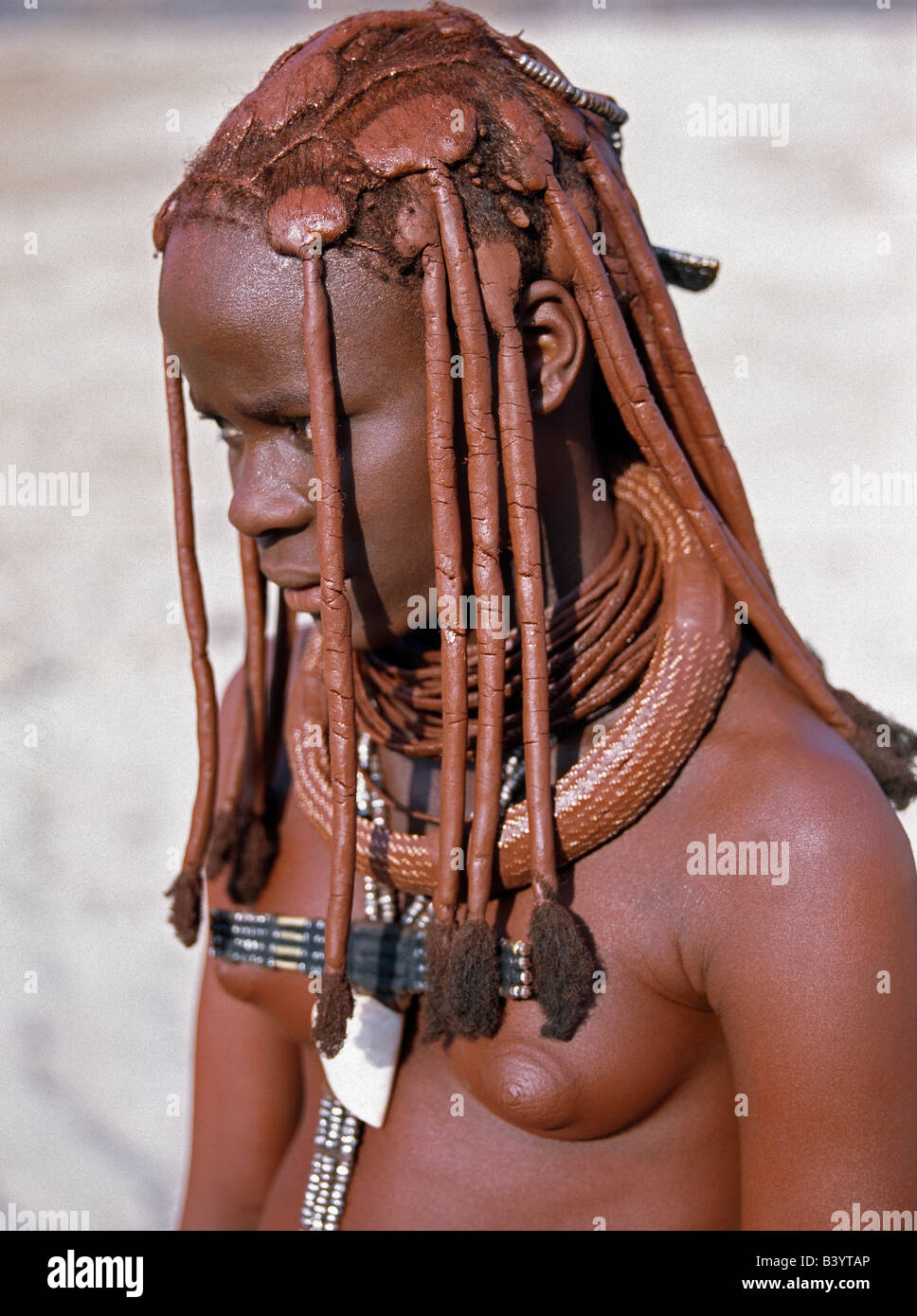 Namibia, Kaokoland, Empembe. A young Himba girl in traditional attire. Her body gleams from a mixture of red ochre, butterfat and herbs. Her long hair is styled in the traditional Himba way. Her large, round white-beaded necklace, called ombwari, is worn by both sexes.The Himba are Herero-speaking Bantu nomads who live in the harsh, dry but starkly beautiful landscape of remote northwest Namibia. Stock Photo