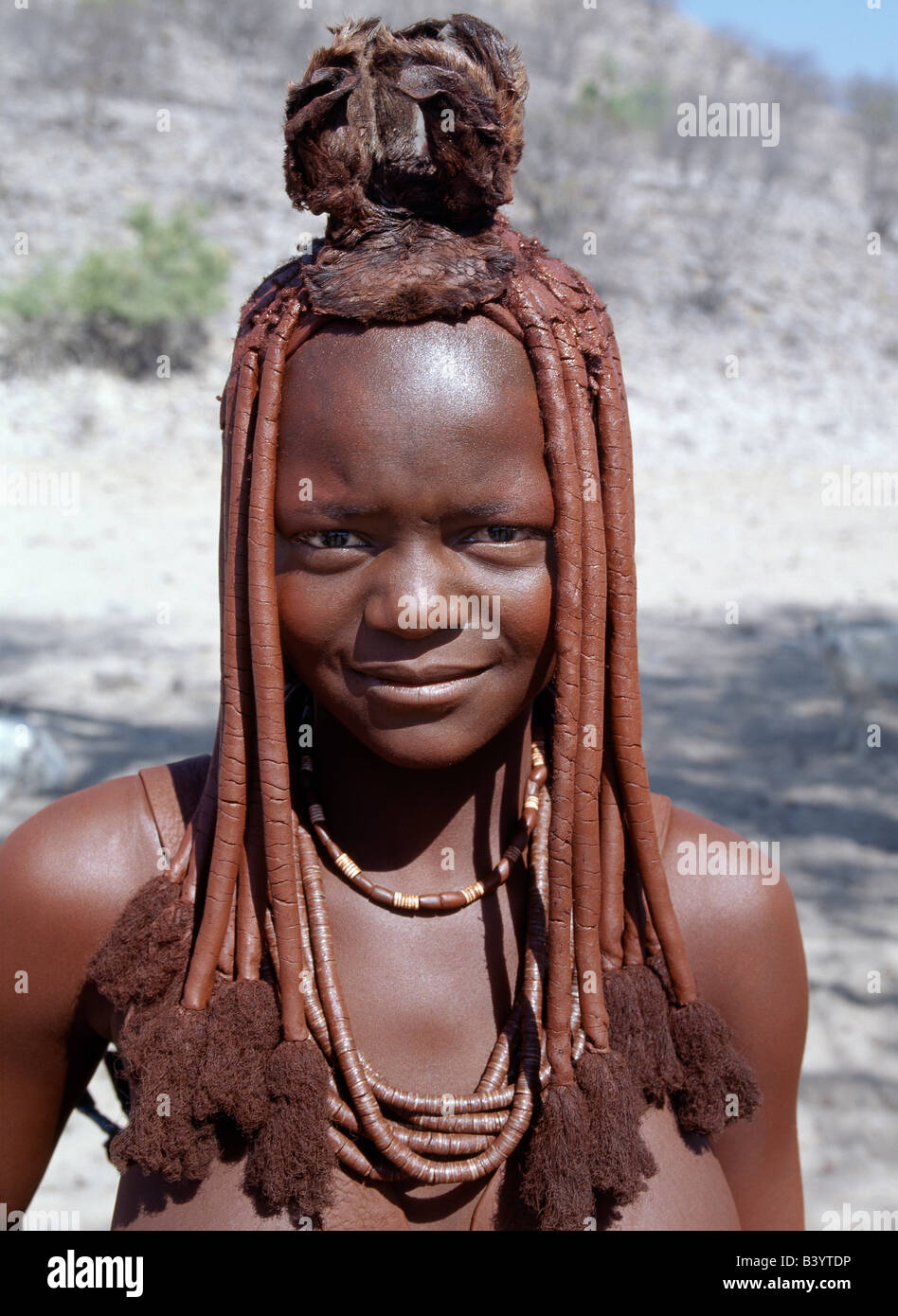 Namibia, Kaokoland, Epupa. A young Himba girl in traditional attire. Her body gleams from a mixture of red ochre, butterfat and herbs. Her hair is styled in the traditional Himba way and is crowned with a headdress made of lambskin, called erembe. Her necklace includes ostrich-shell beads. The Himba are Herero-speaking Bantu nomads who live in the harsh, dry but starkly beautiful landscape of remote northwest Namibia. Stock Photo