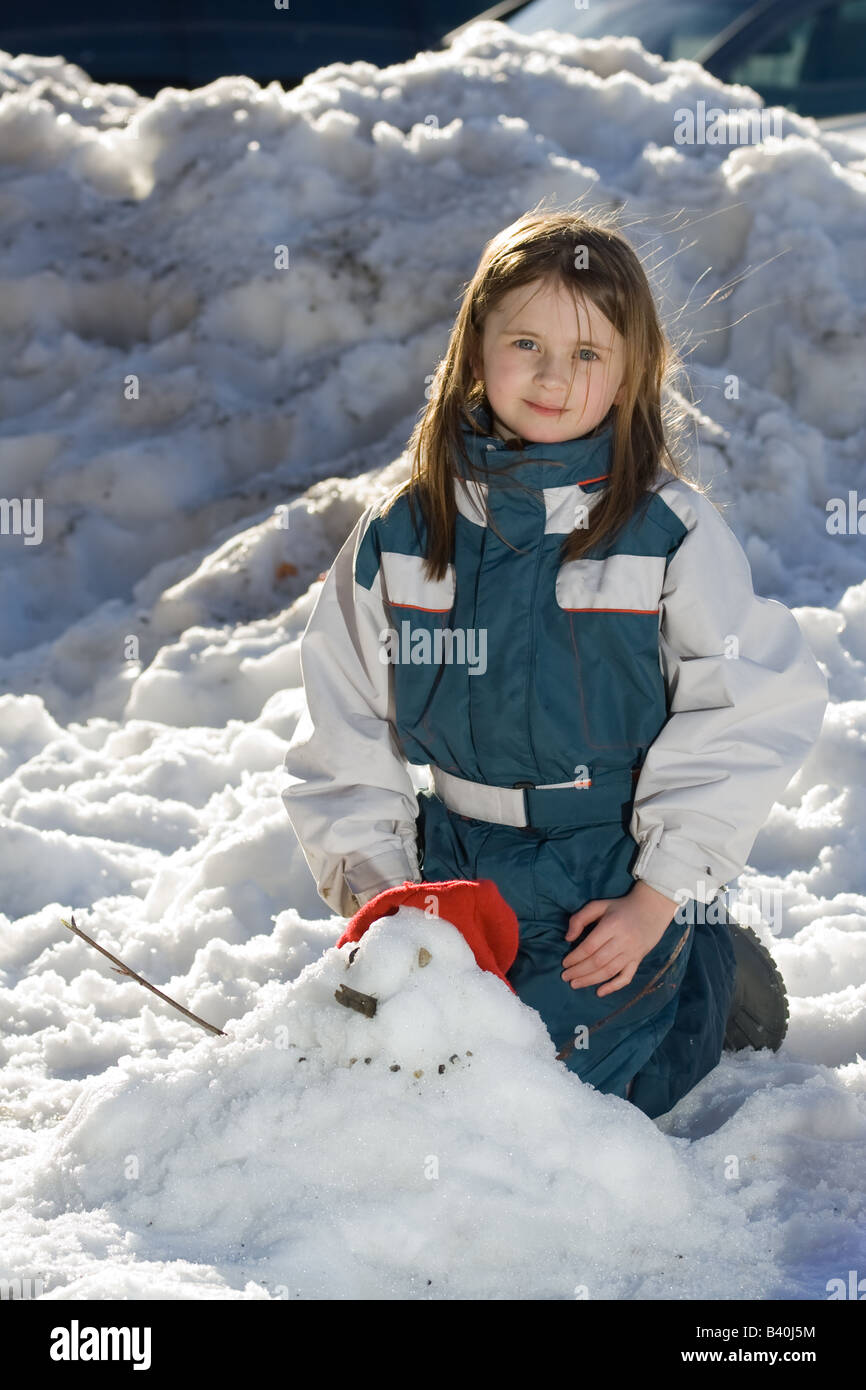 Portrait of a girl behind a squat snowman she has made with a red cap and stick arms She is wearing a blue and white ski outfit. Stock Photo