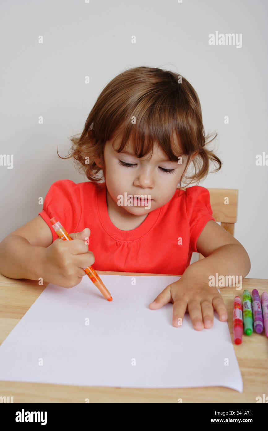 A two years old toddler girl is drawing awkwardly holding an orange colored crayon. Stock Photo