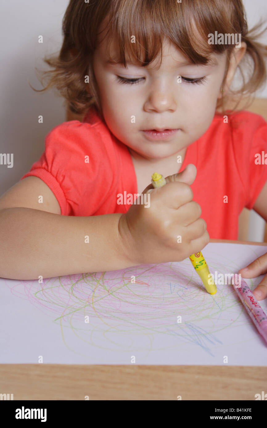 A two years old toddler girl is drawing awkwardly holding an yellow colored crayon. Stock Photo