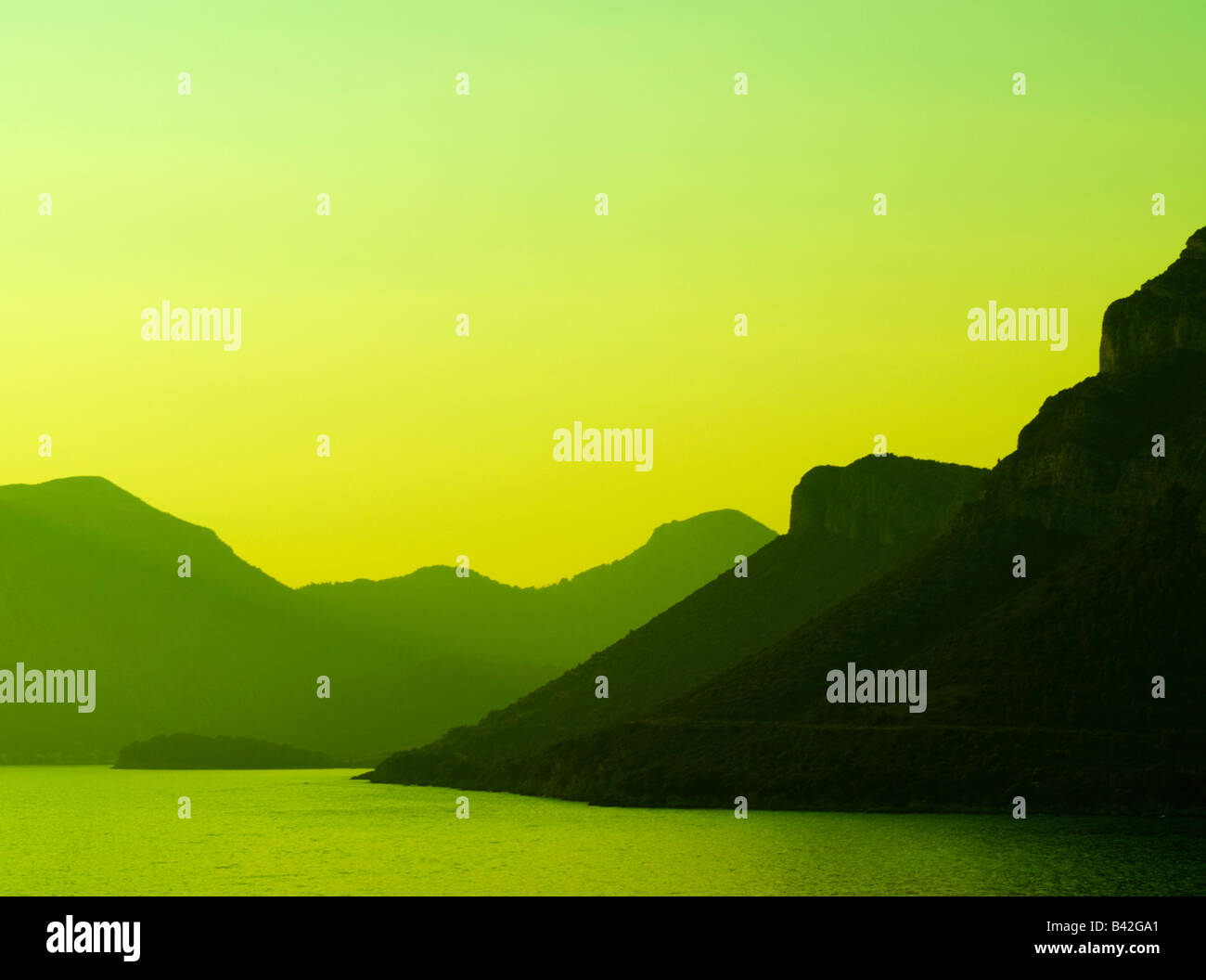 Heavy dusk. Fading heavy green light over mountains that get lost in calm ocean at their feeet. Stock Photo
