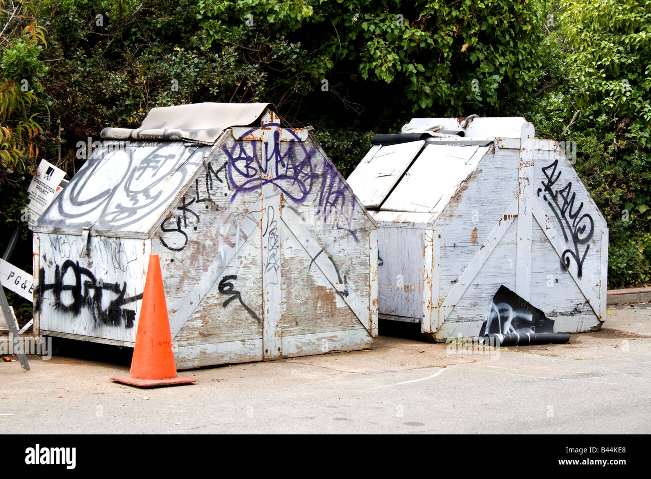 Two graffitti filled industrial trash receptacles with an orange warning cone in front Stock Photo