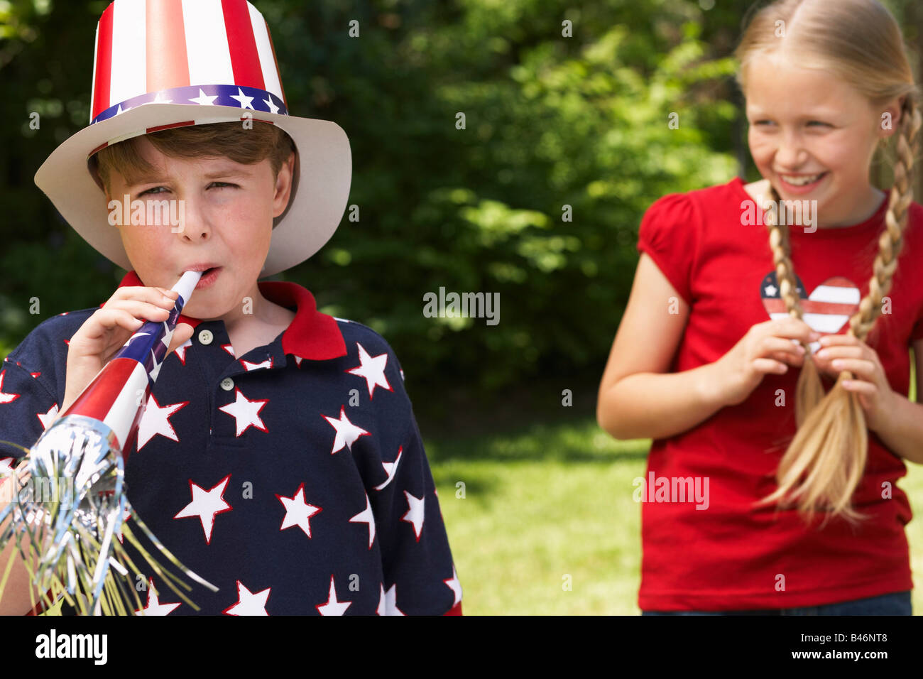 Portrait of Boy Wearing Stars and Stripes Top and Hat, Blowing Noisemaker Horn, Girl Watching Stock Photo
