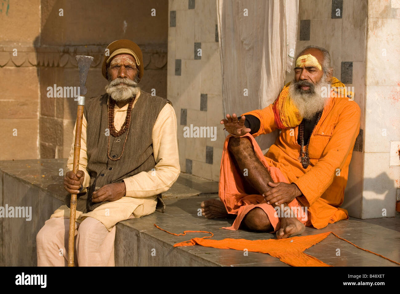 A bearded sadhu sits in contemplation by a temple in the city of Varanasi, India. A man with a staff sits next to him. Stock Photo