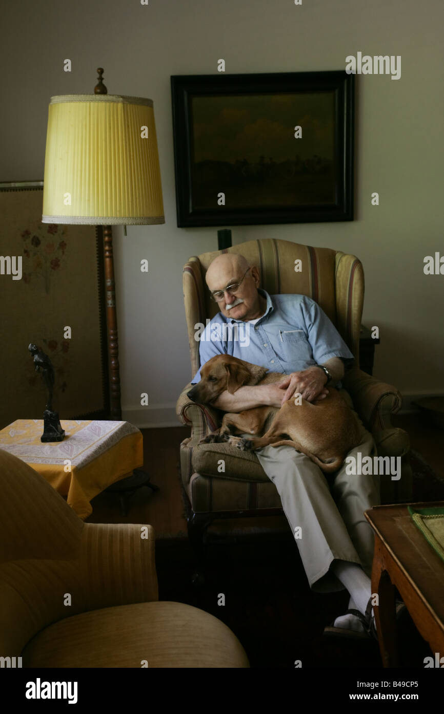 An old man comforts a dog he is holding on his lap inside a dark room with antique furniture. Stock Photo
