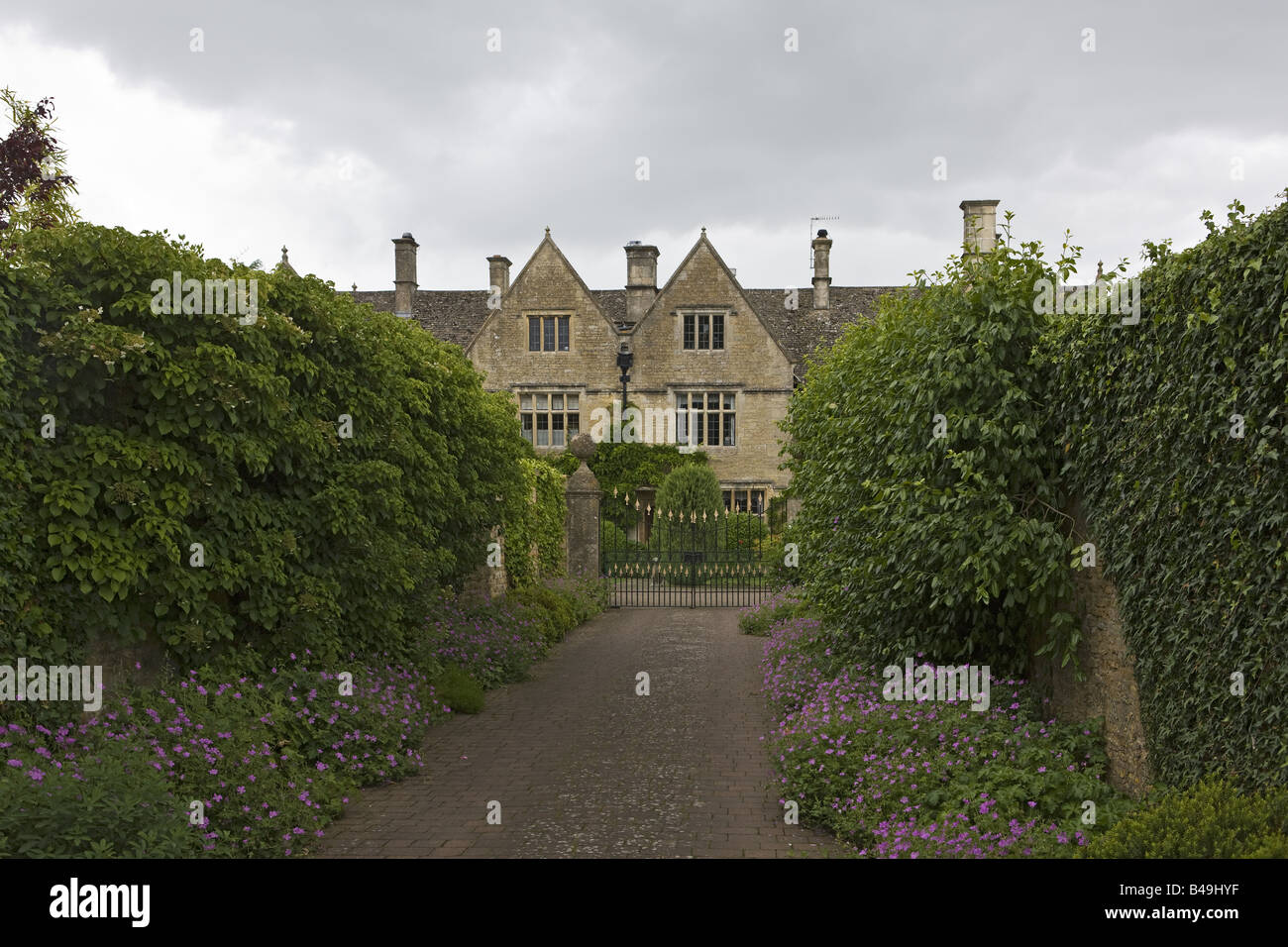 Cotswold's Family Mansion House Viewed Down Shrubbery & Flower Lined Drive Through Gates Stock Photo