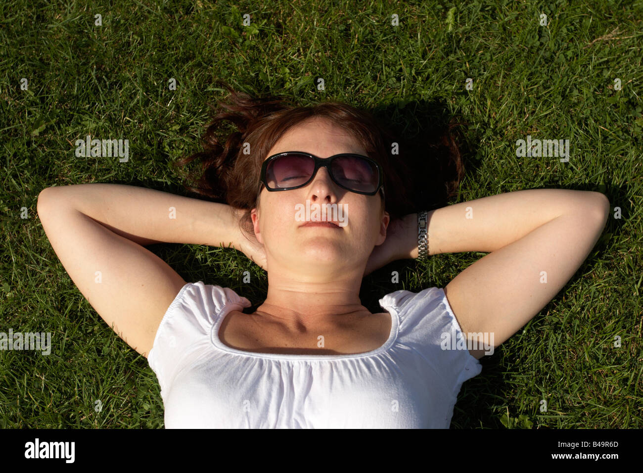 Woman outdoors laying on her back sunbathing on grass wearing sunglasses. Overhead view. Stock Photo