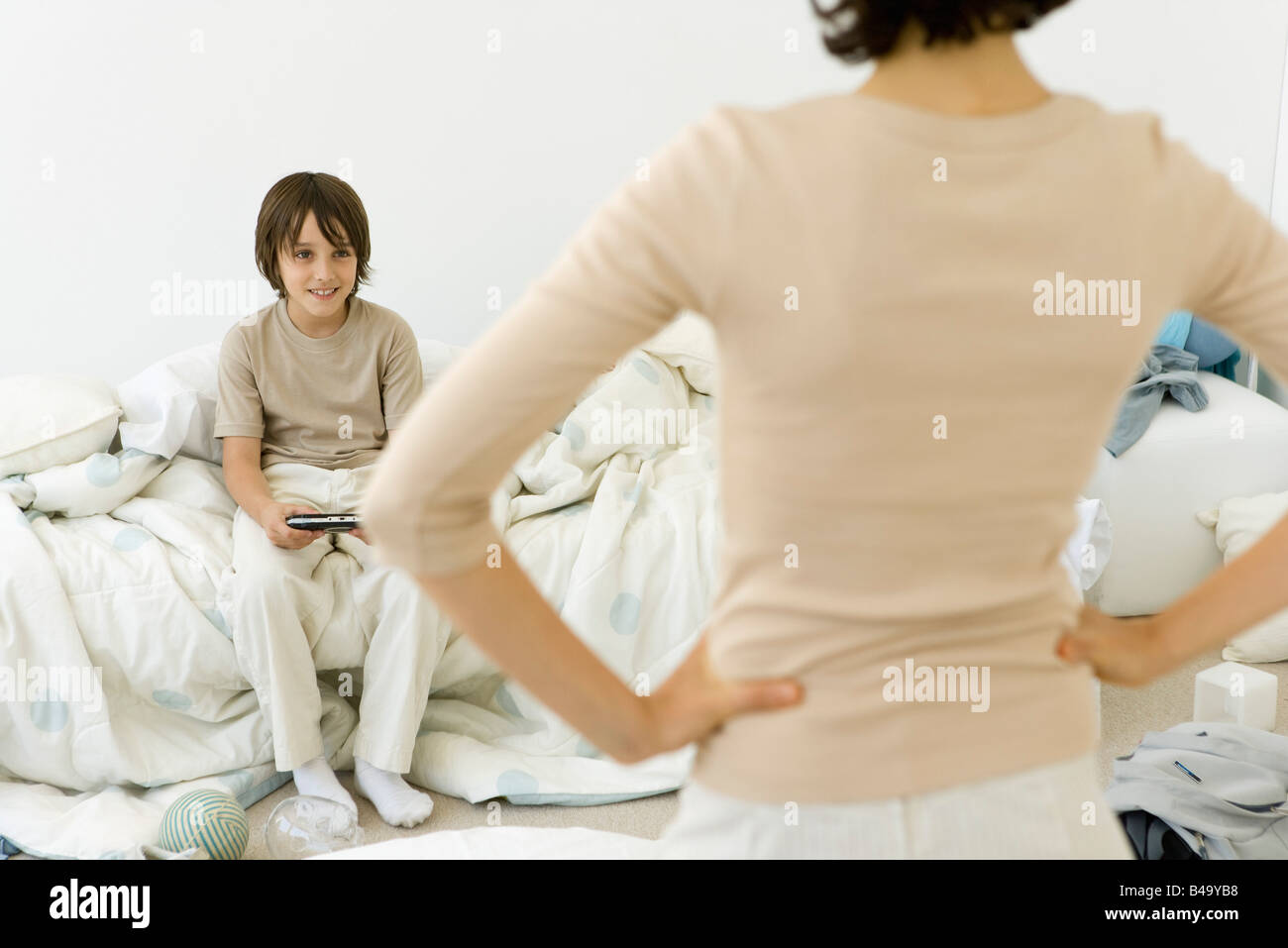Boy playing handheld video game in messy bedroom, mother standing in foreground with hands on hips Stock Photo