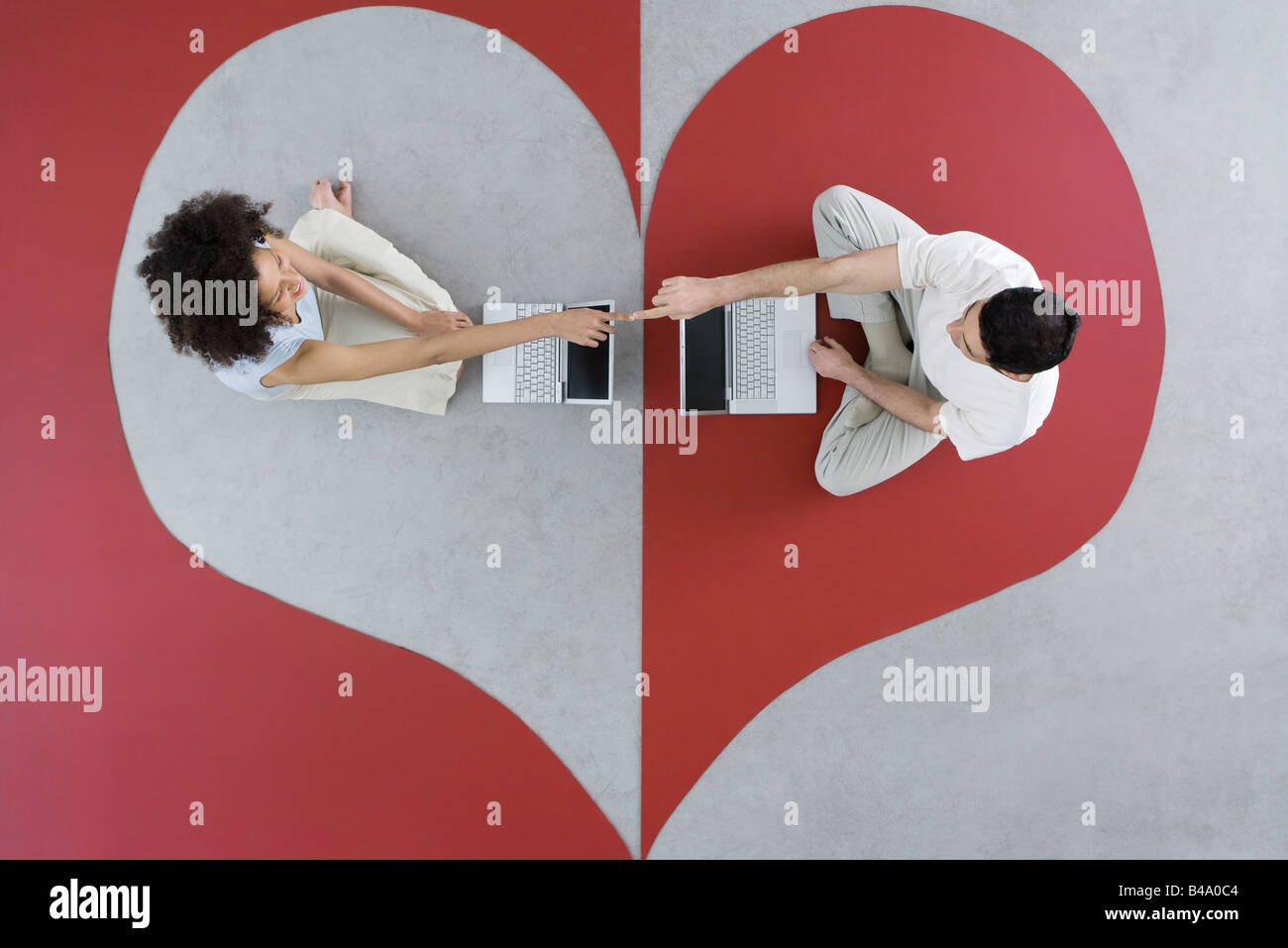 Couple sitting face to face with laptop computers on heart shape, touching fingers, overhead view Stock Photo