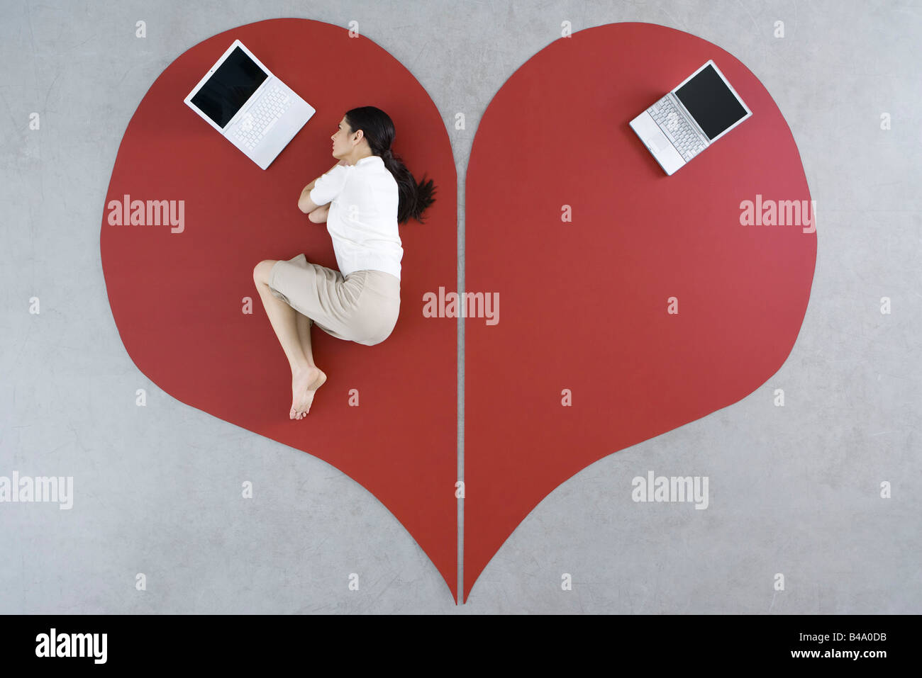 Woman lying on the ground with laptop on large broken heart, abandoned laptop behind her Stock Photo