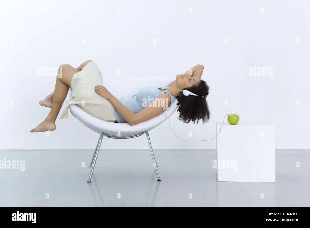 Woman reclining in chair, listening to headphones connected to apple, side view Stock Photo
