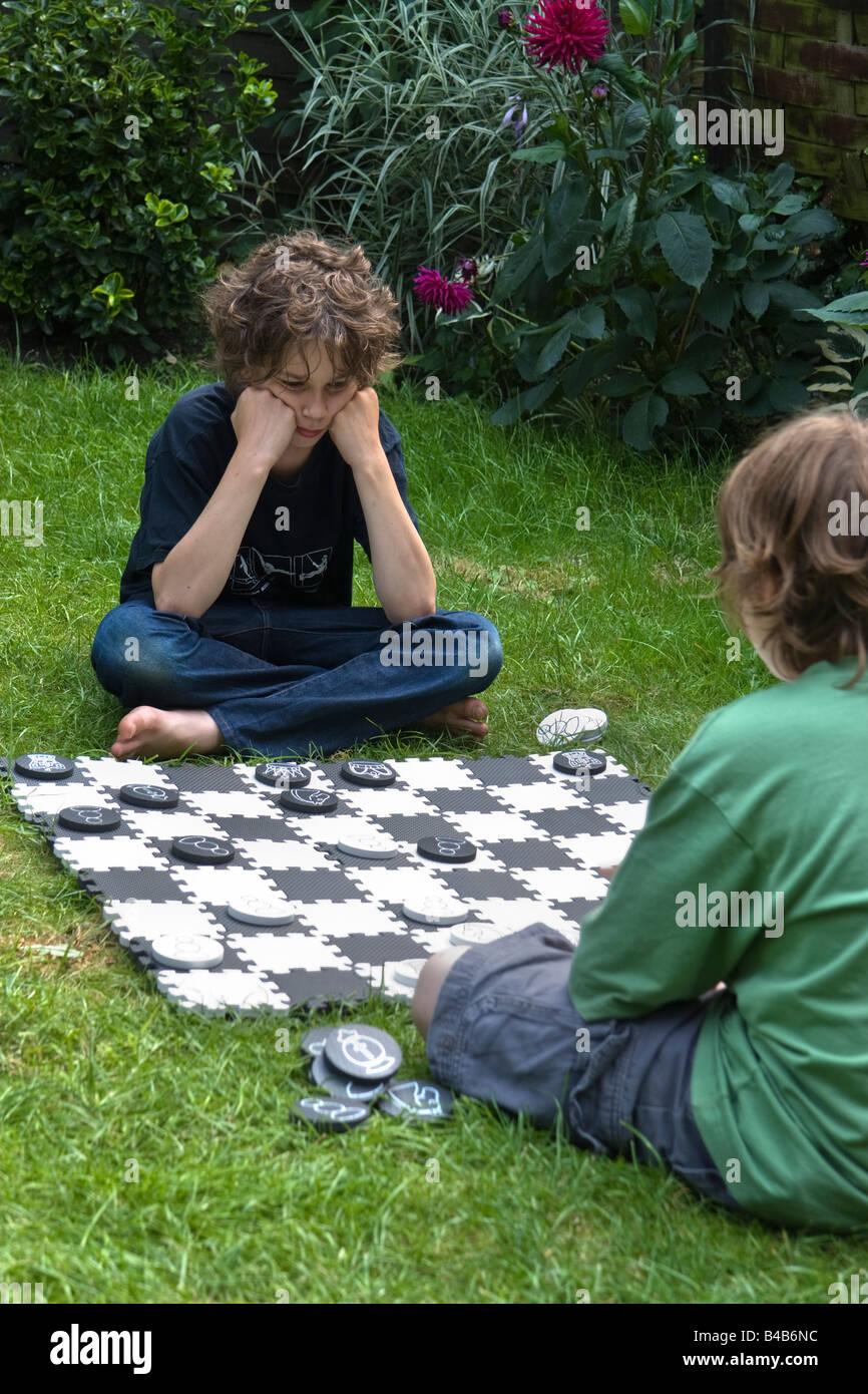 Two boys sitting on the grass in a garden playing chess on an outsize chessboard Stock Photo