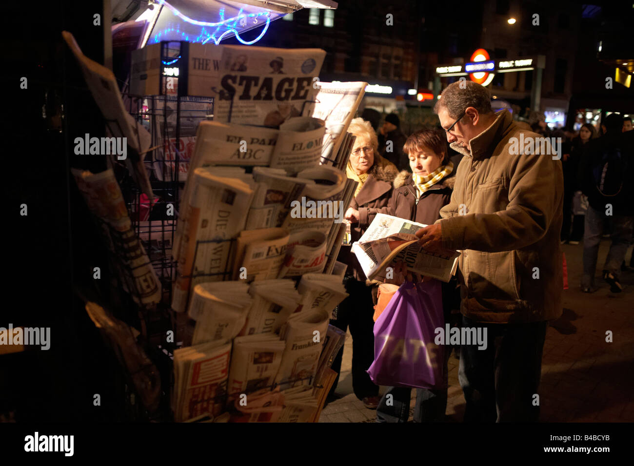 Foreign visitors to Theatreland in London's West End read newspapers at a kiosk lit by spotlights on Tottenham Court Road Stock Photo
