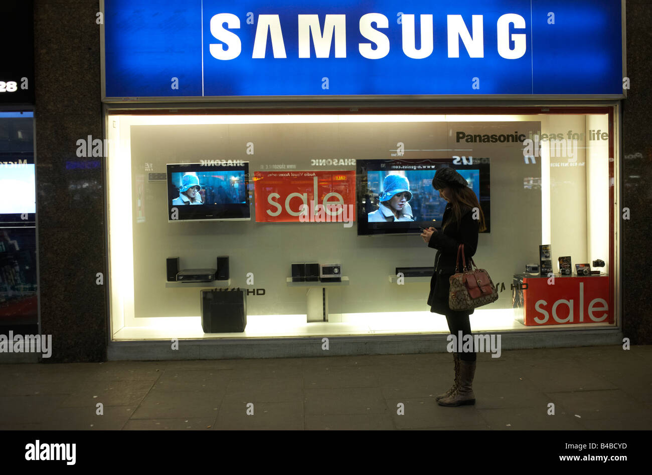 A young lady texts a friend outside an electronics shop in London's Tottenham Court Road where TVs show the King Kong film. Stock Photo