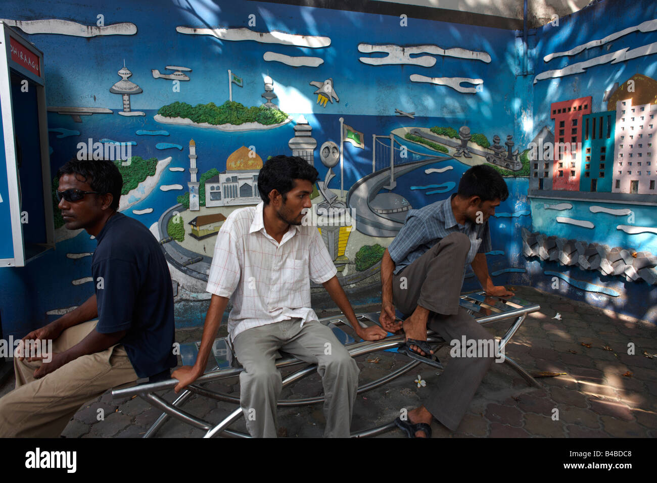 In the shade of mid-day heat near a mural  Maldivian youths sit about on a park bench in the Maldives capital Malé Stock Photo