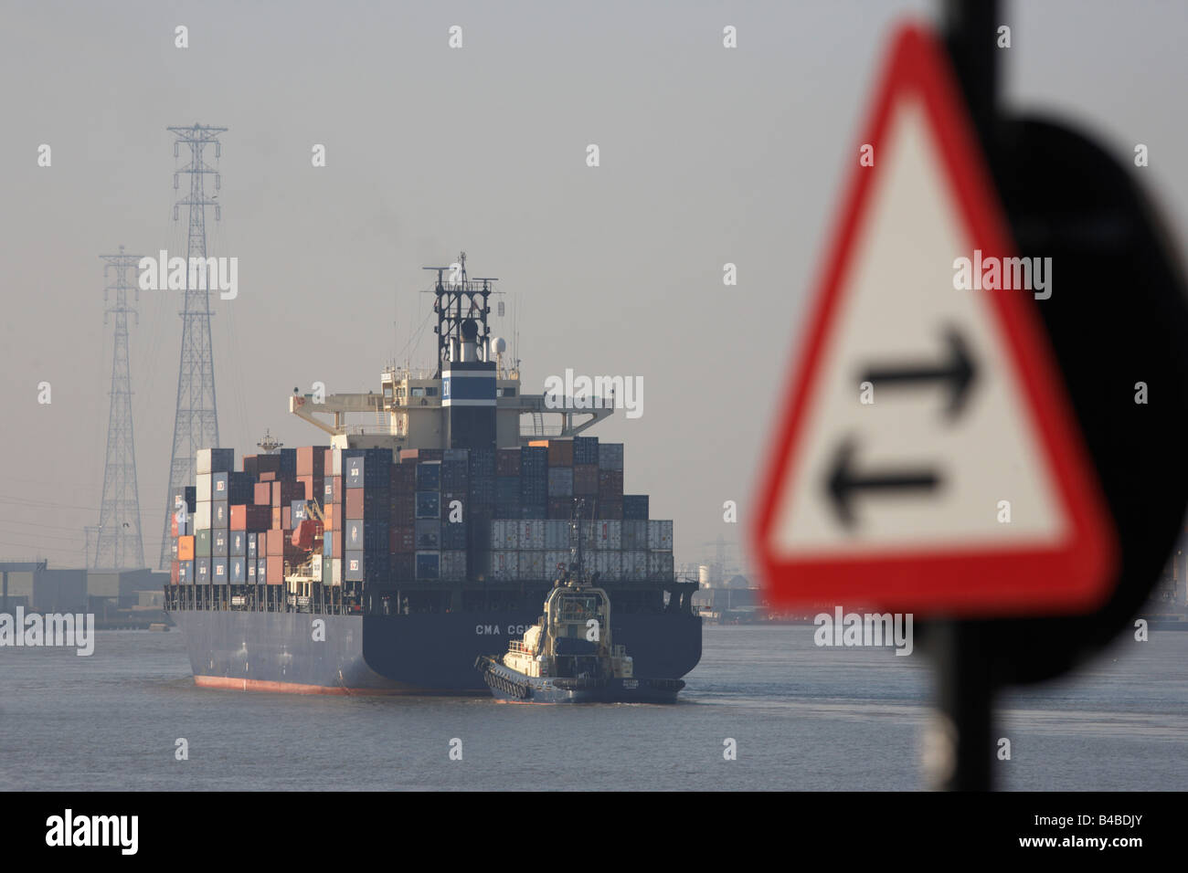 Past a two-way sign, a giant cargo container ship is guided by tug, easing upstream on the River Thames towards Tilbury Docks Stock Photo