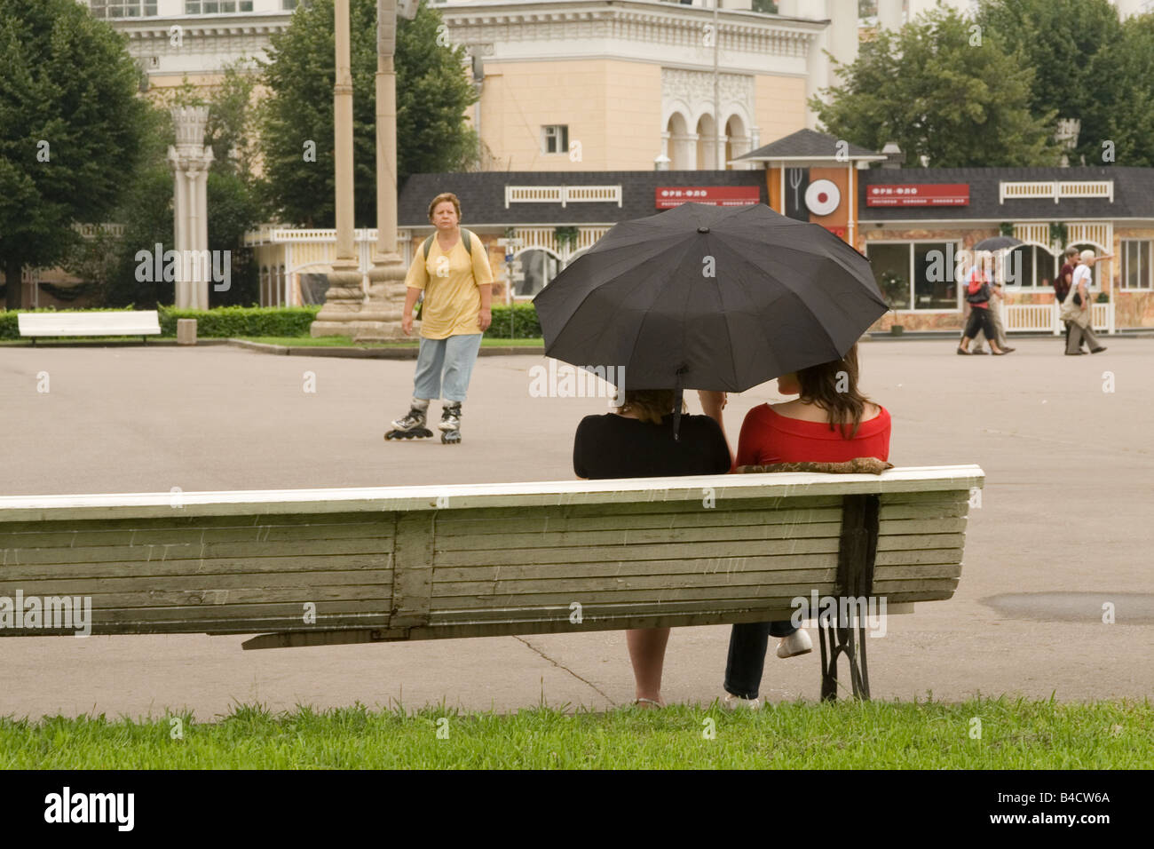 Friends sit under umbrella on a bench at the All-Russia Exhibition Centre. Stock Photo