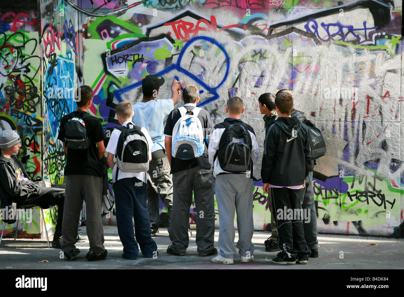 Group of young boys men teenagers hoodies delinquents skaters watch a graffiti artist at work spraying graffiti on a wall London Stock Photo