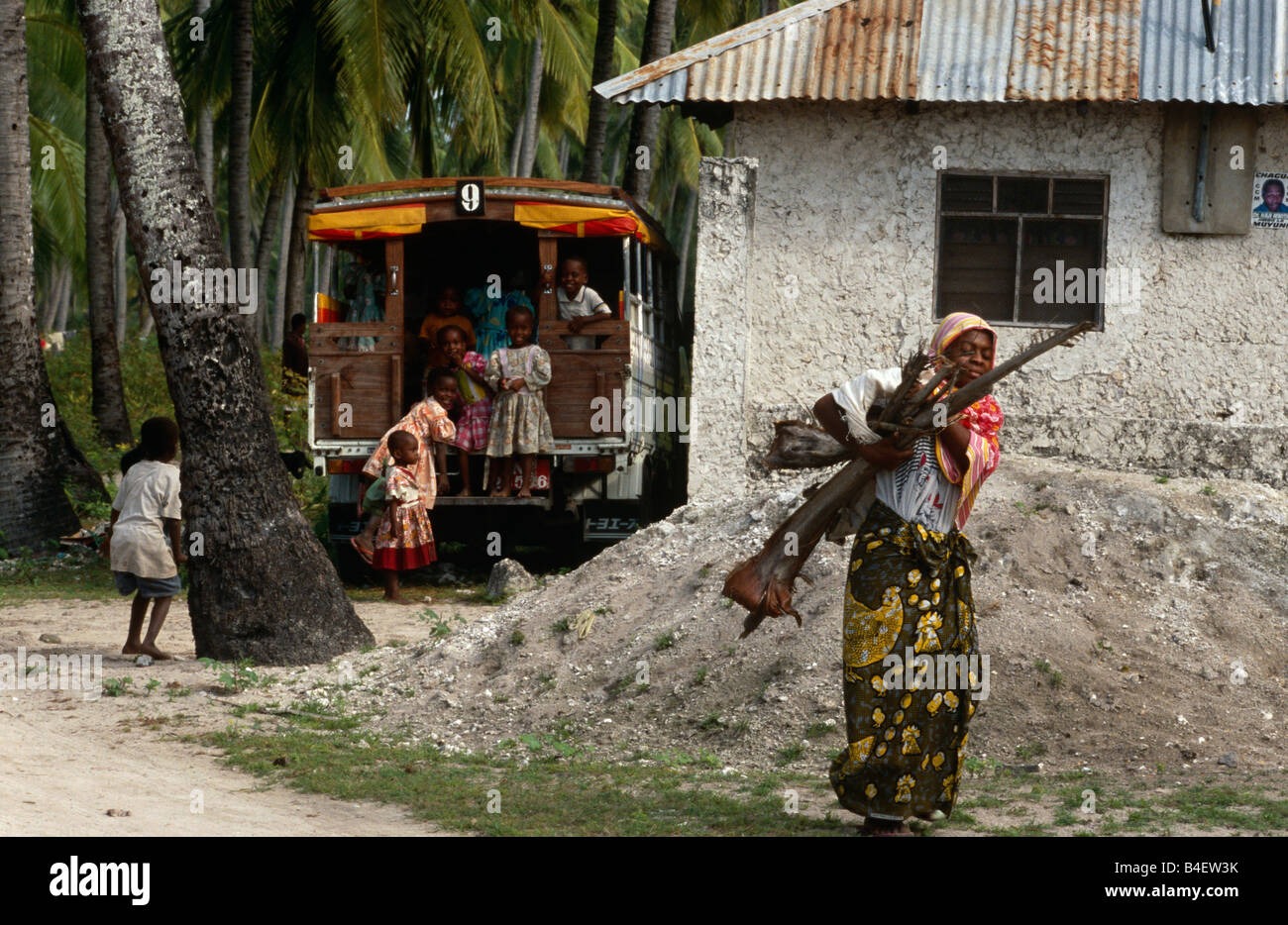 Woman collecting firewood, children playing on bus in background in village. Zanzibar. Stock Photo