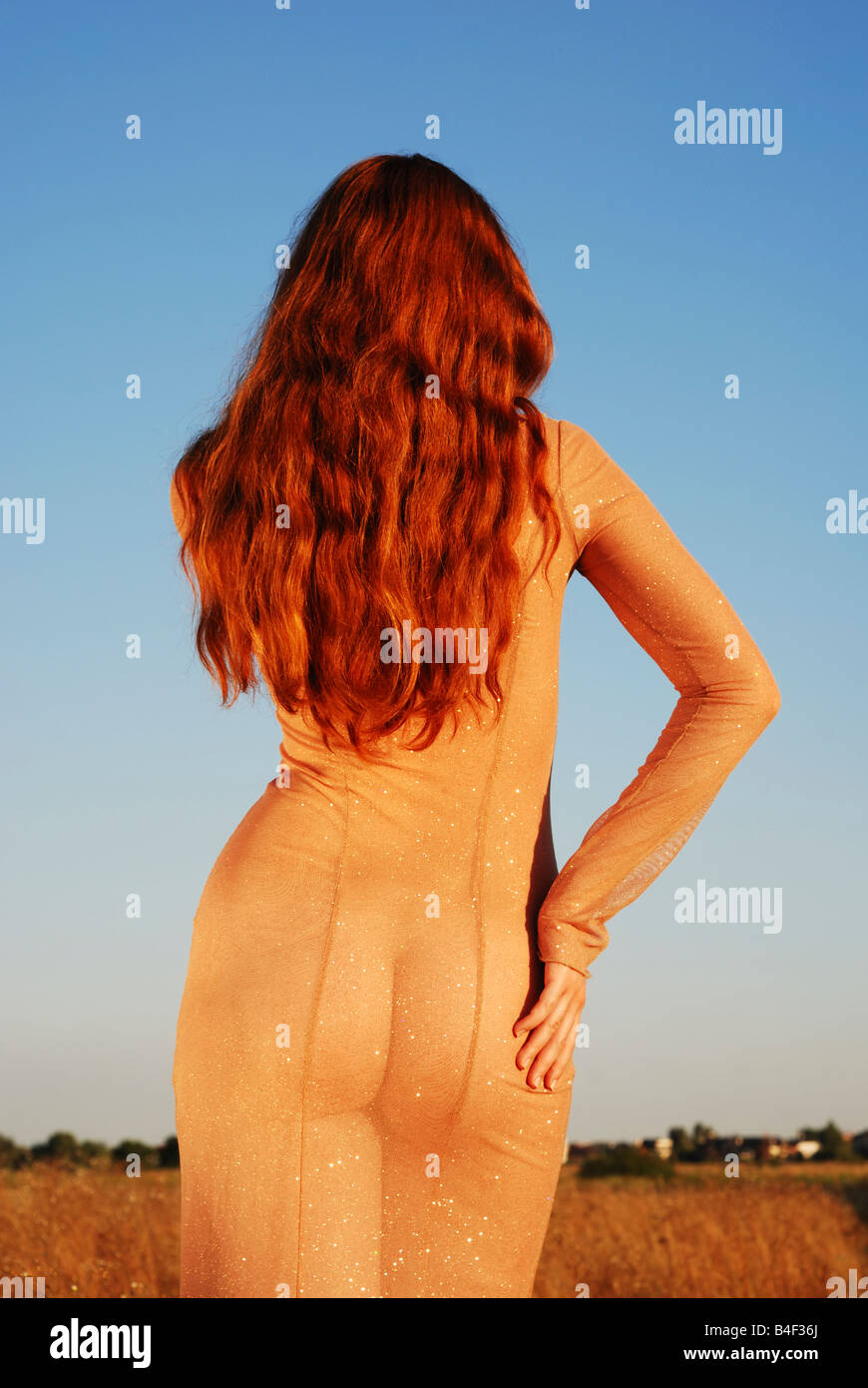 Long-haired girl in transparent beige dress stands back against sky and field, right hand on hip, warm tint of summer sunset Stock Photo