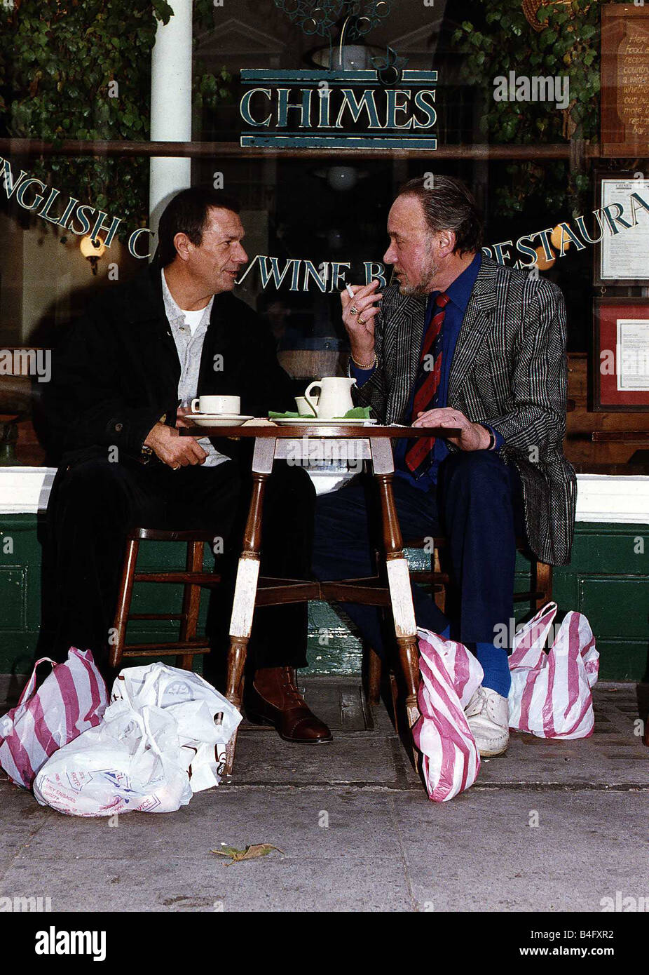 Don Henderson Actor Michael Elphick actor sitting at pavement cafe table with shopping bags talking and smoking a cigarette Stock Photo