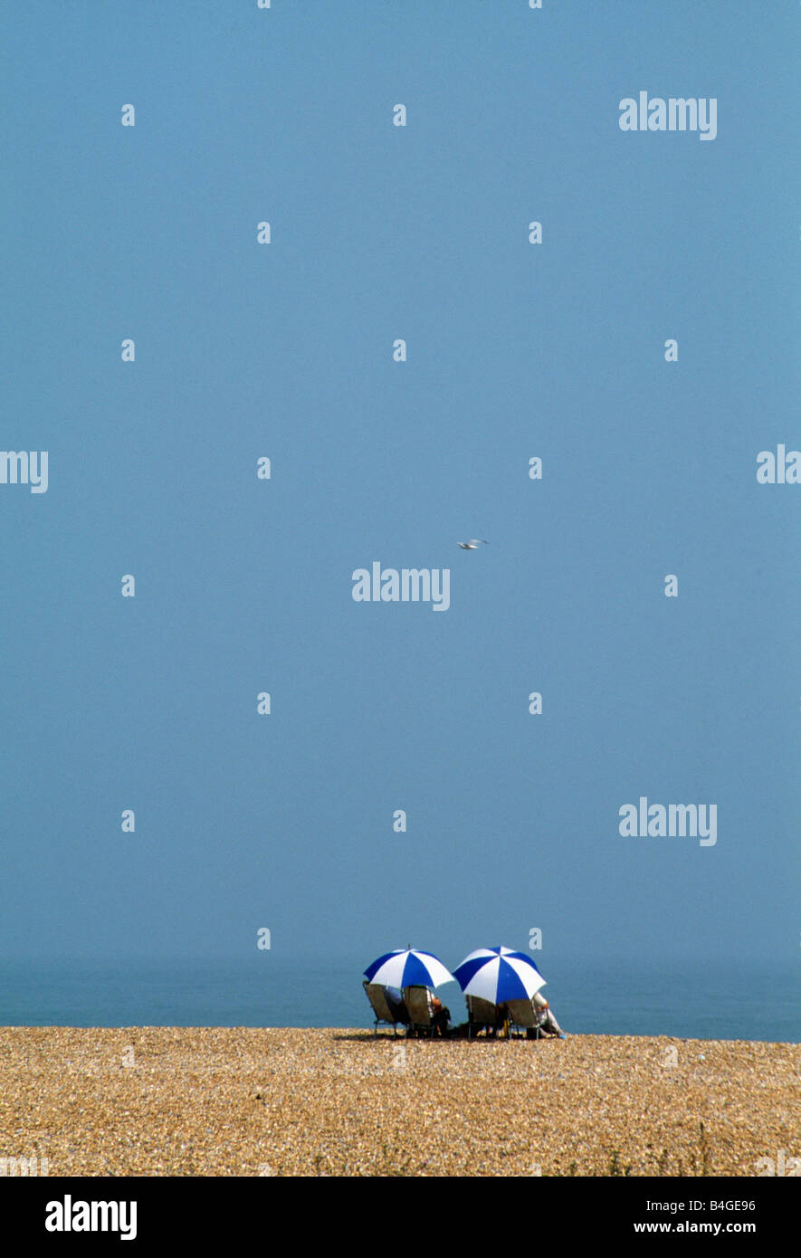 Blue sky above couple sitting beneath blue white umbrellas on the beach at Deal in Kent Stock Photo