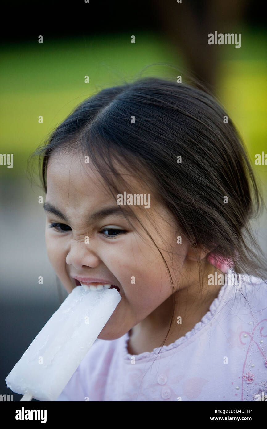 Small half-Thai girl is eating an iced lolly in a play area one evening but the ice is so cold it makes her pull a face Stock Photo