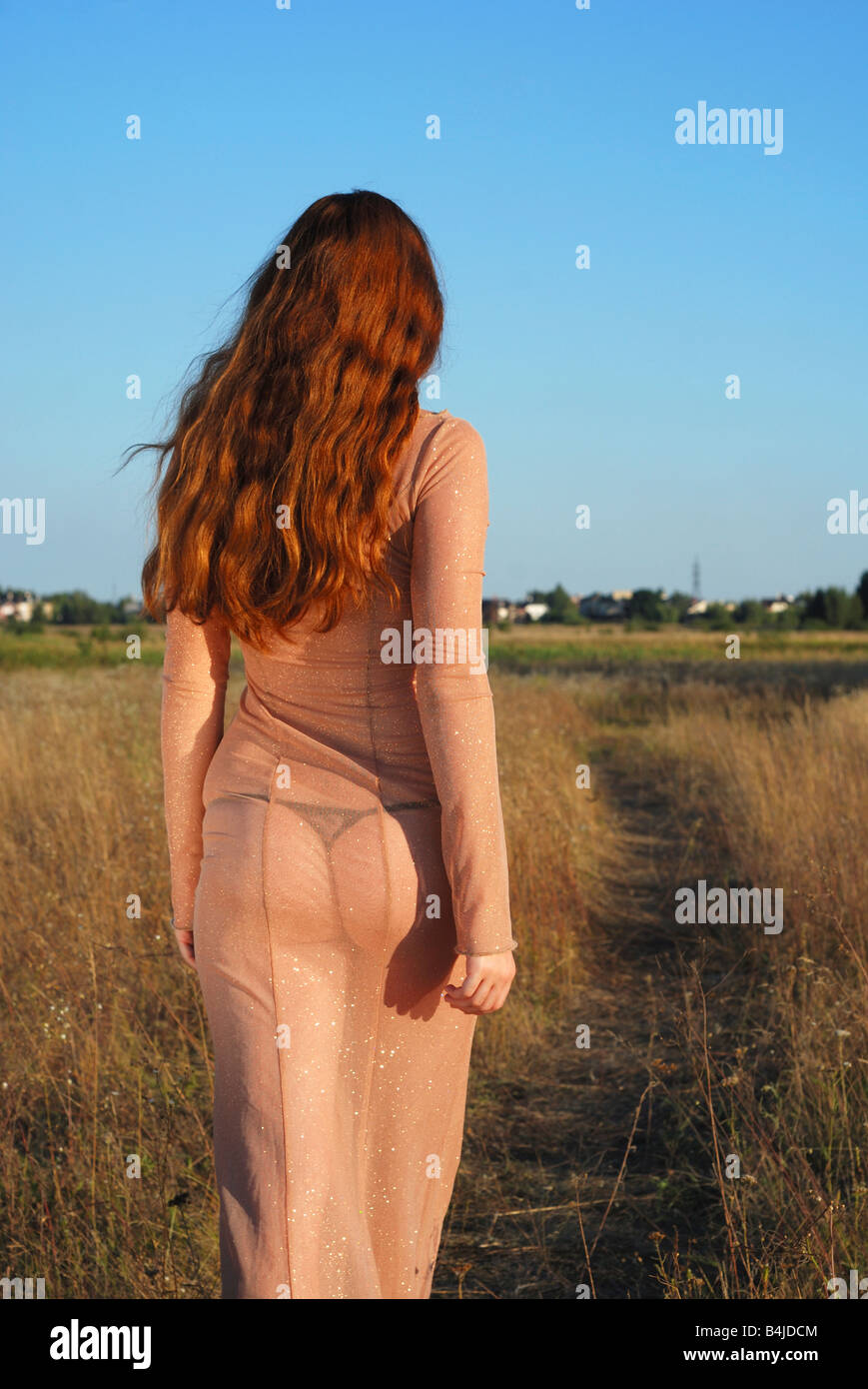 Long-haired girl going on path in transparent flesh-colored dress against blue sky and meadow dried, warm tint of summer sunset Stock Photo