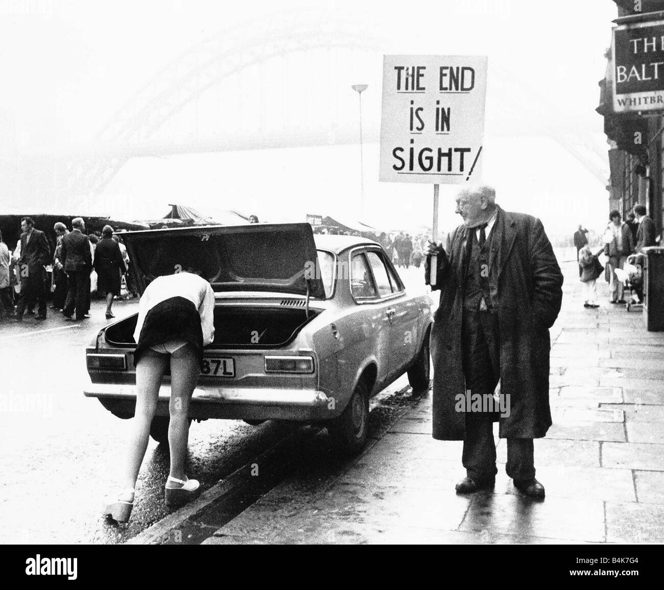 A woman is bending down leaning into the boot of her car revealing her white knickers as a old man holding a banner saying THE END IS IN SIGHT turns and looks Stock Photo