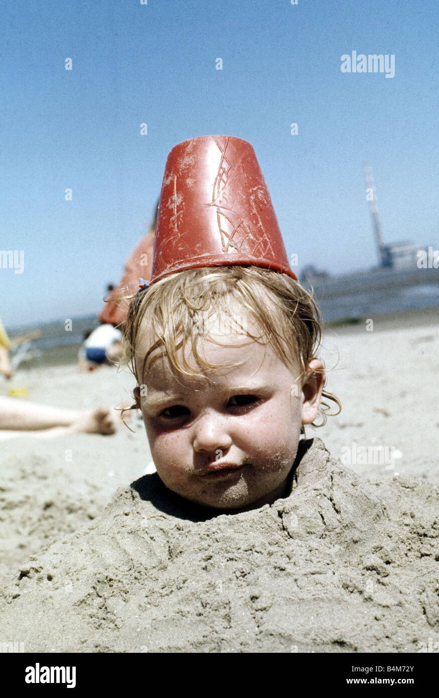 Summer holidays July 1976 Boy buried in sand with red plastic Bucket on his head Stock Photo
