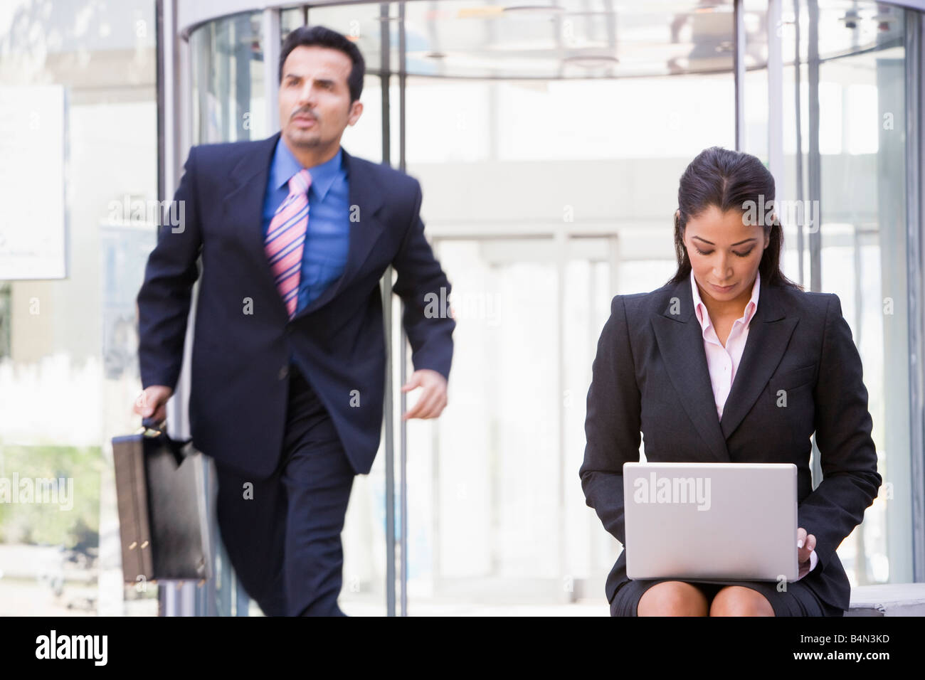 Businesswoman outdoors in front of building using laptop with businessman in background (high key/blur) Stock Photo