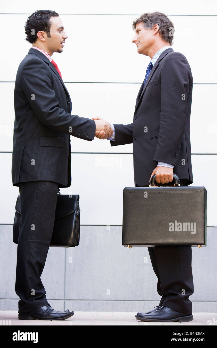 Two businessmen shaking hands outdoors holding briefcases and smiling Stock Photo