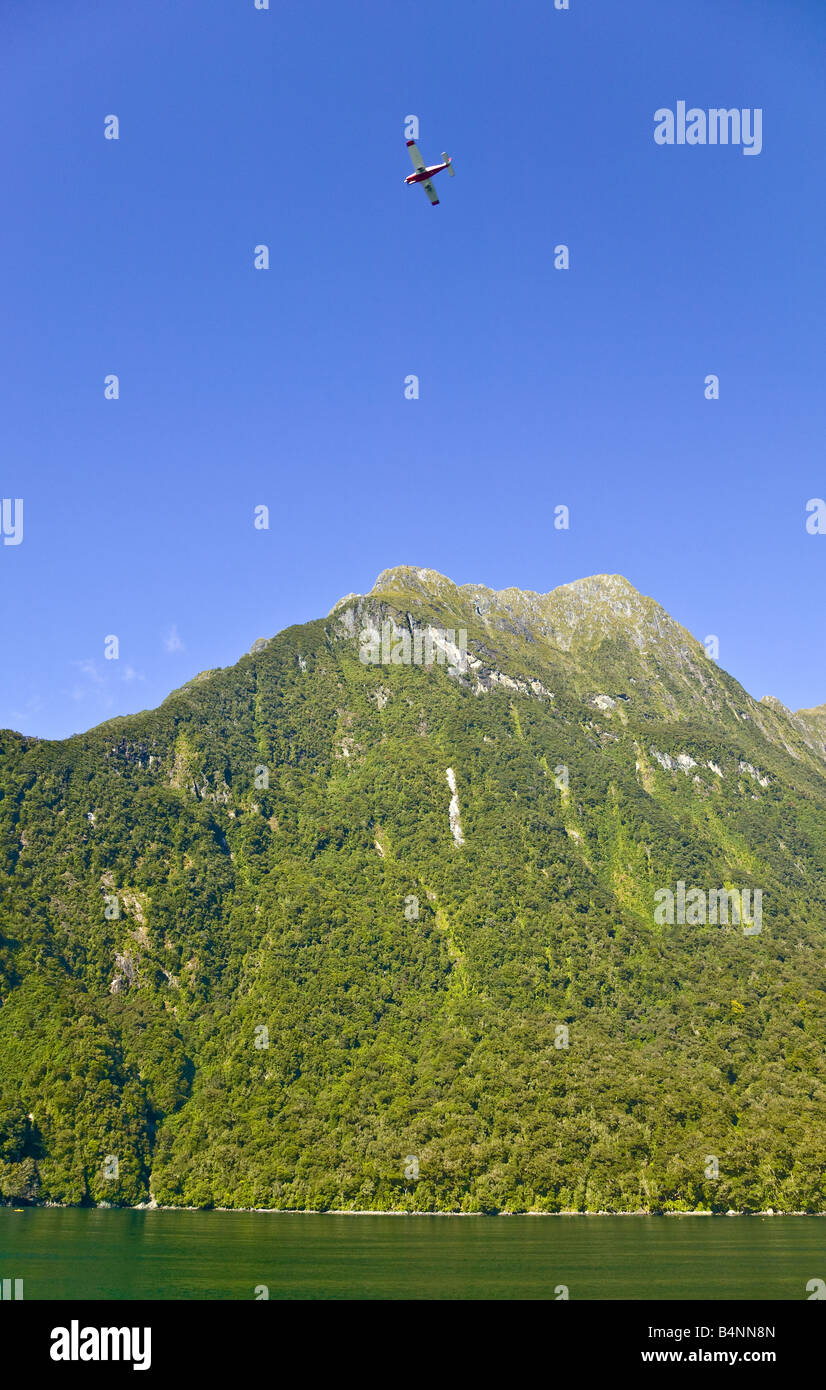 Forest covered mountain summit above the south bank of Milford Sount with a tourist plane flying in a blue sky abouve it. Stock Photo