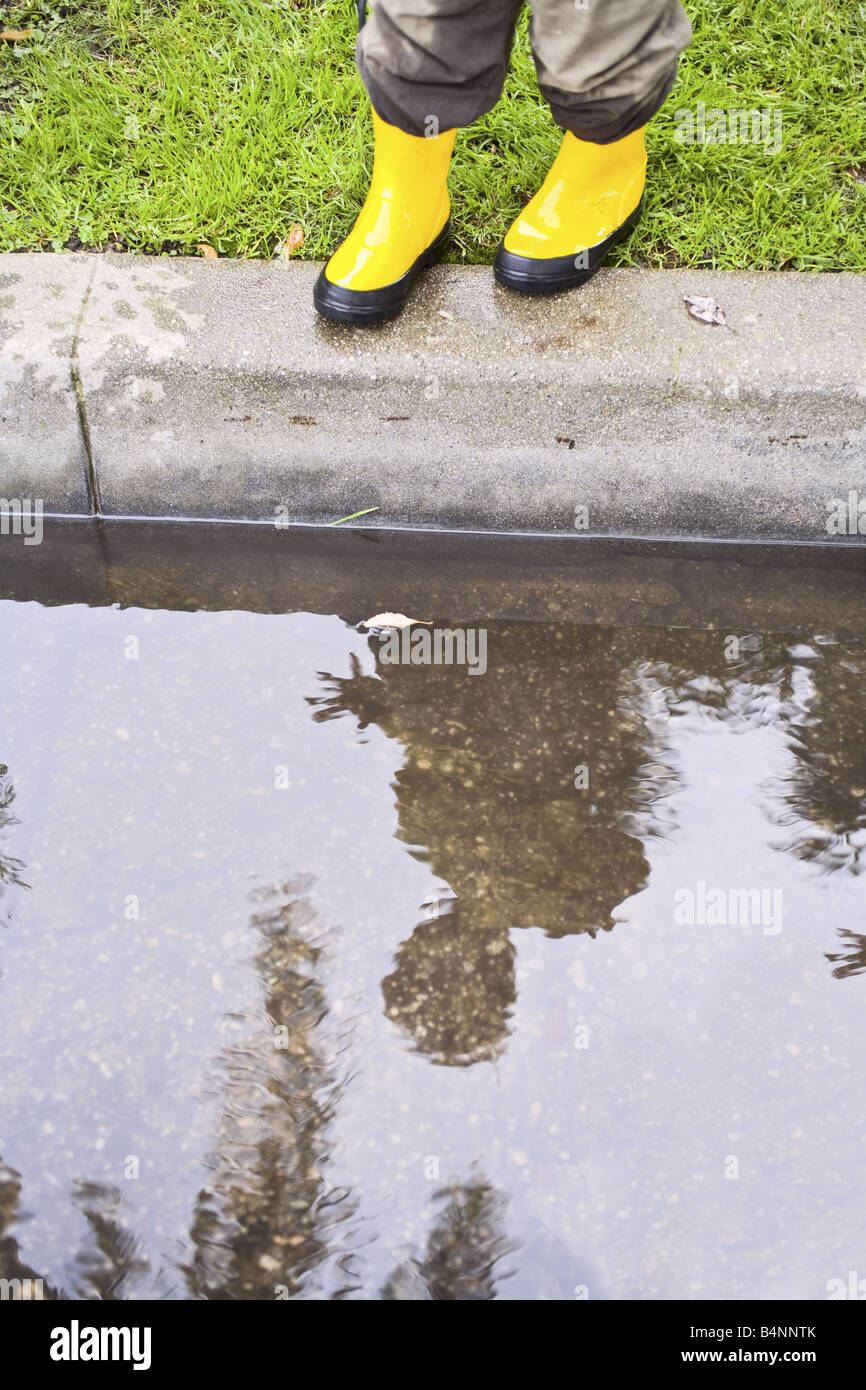 A child standing over a puddle and looking at his reflection in yellow rain boots. Stock Photo
