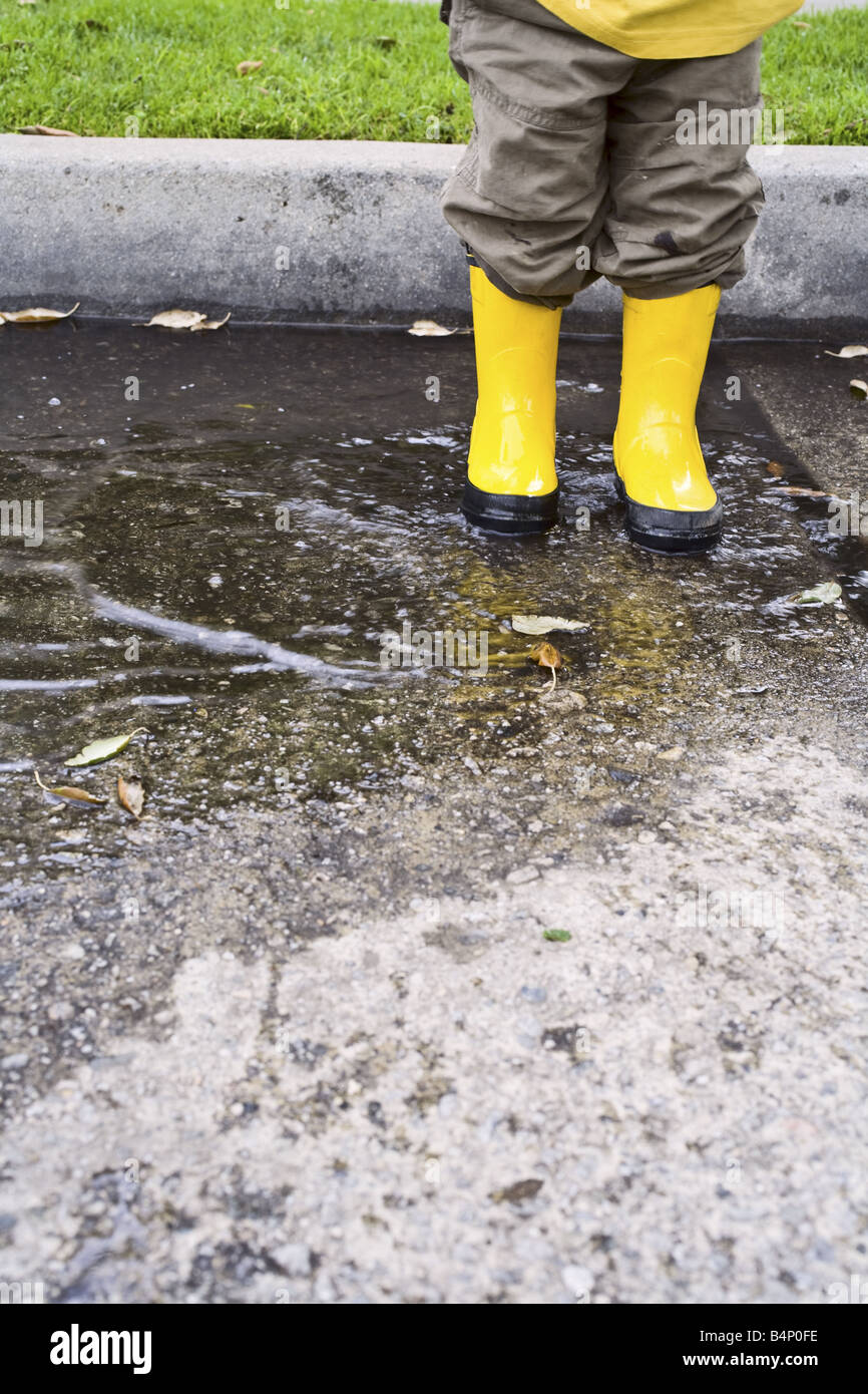A toddler boy stands in a puddle with yellow rain boots on. Stock Photo