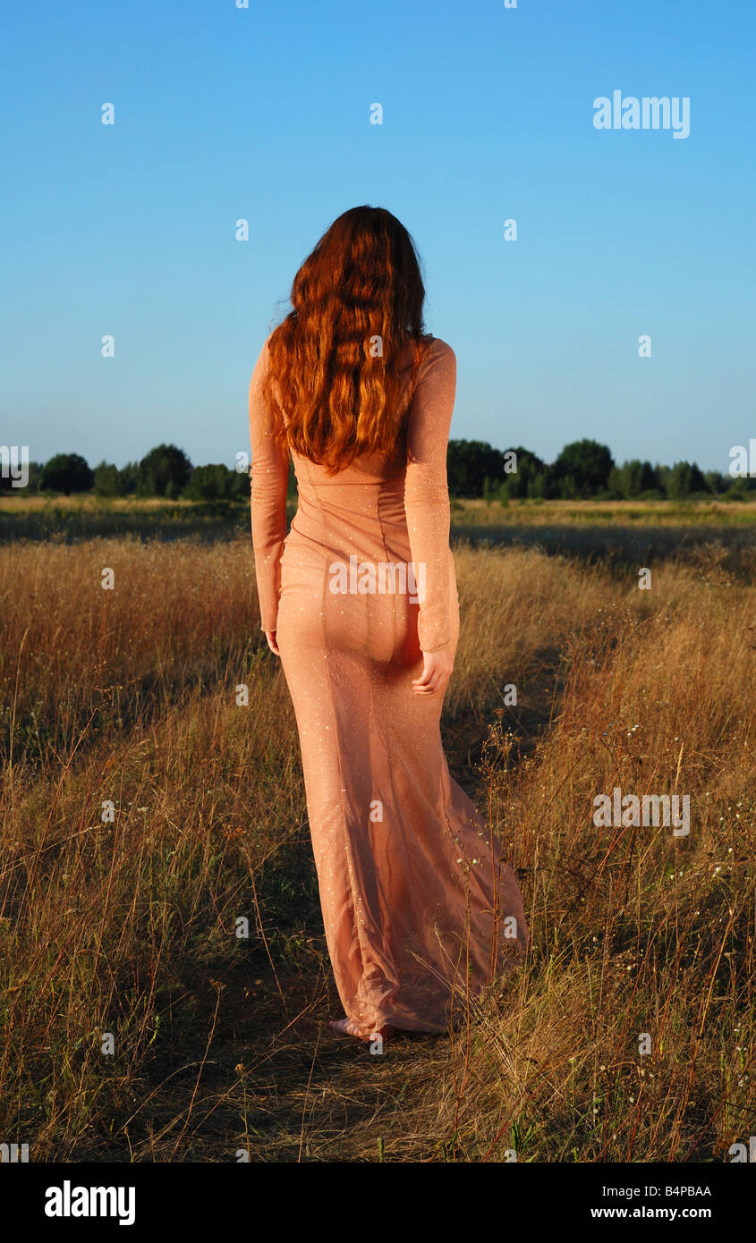 Long-haired girl going on path in transparent flesh-colored dress against blue sky and meadow dried, warm tint of summer sunset Stock Photo