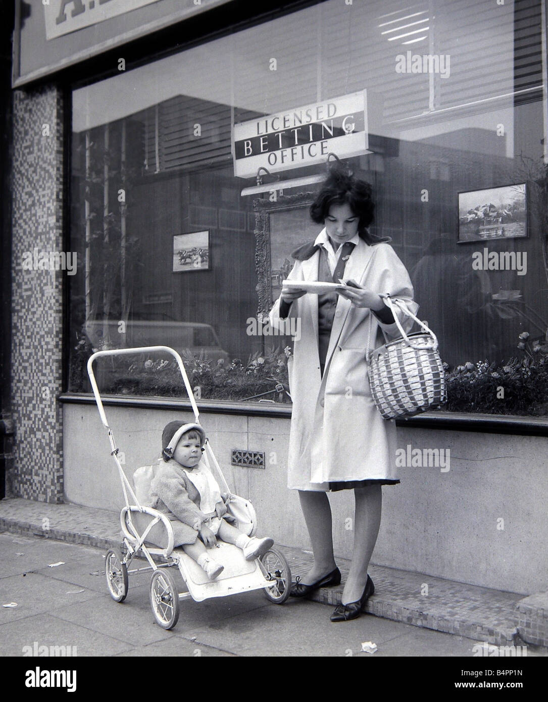 A woman punter studies the form guide outside A E Fane and Co betting shop in islington London as her baby daughter waits patiently in her buggy May 1961 Stock Photo