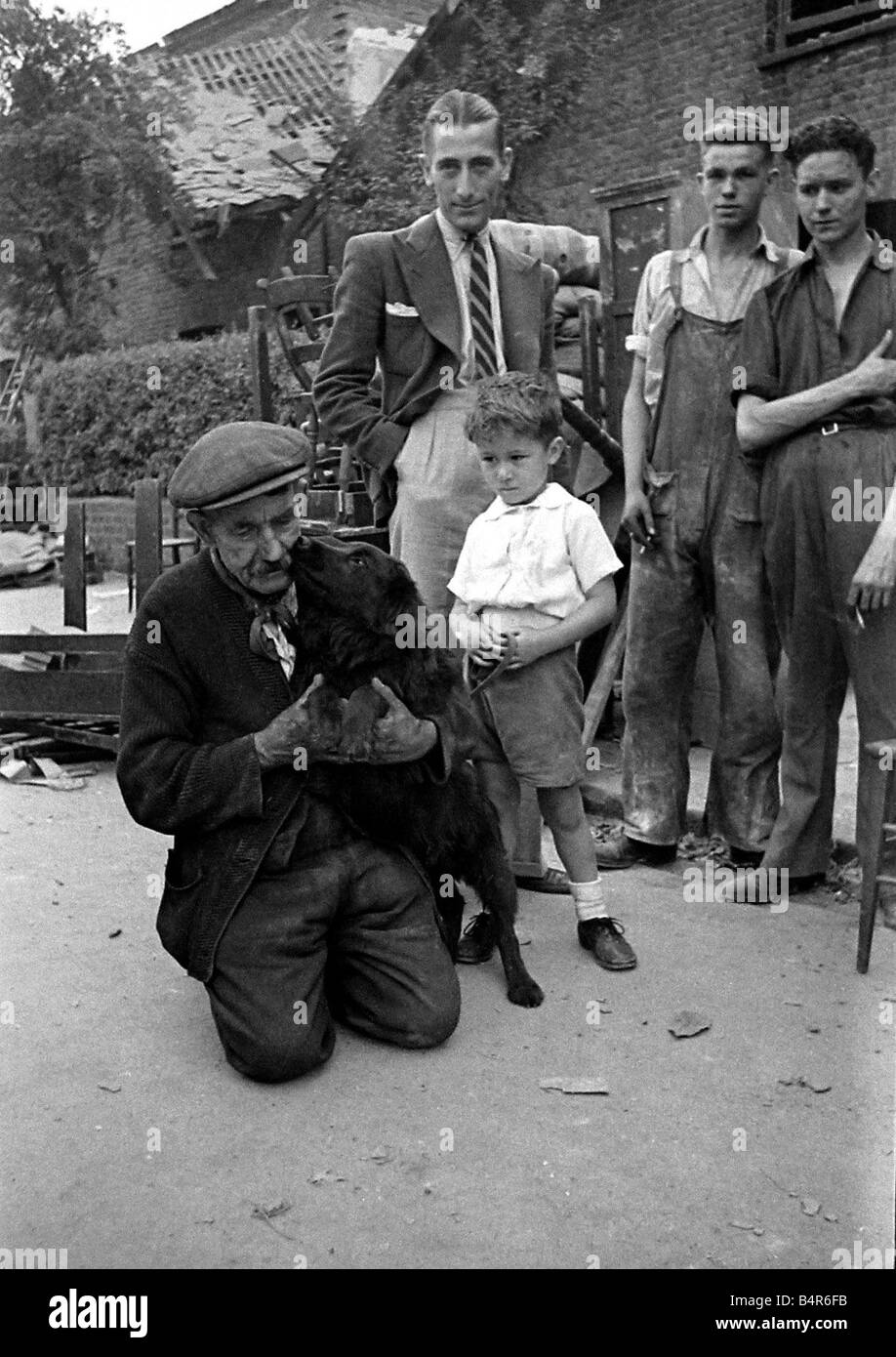 A old man holds his dog as it licks his face watched by a young boy and three men in bomb damaged St Mary s Road Edmonton following air raids in London July 1944 Stock Photo
