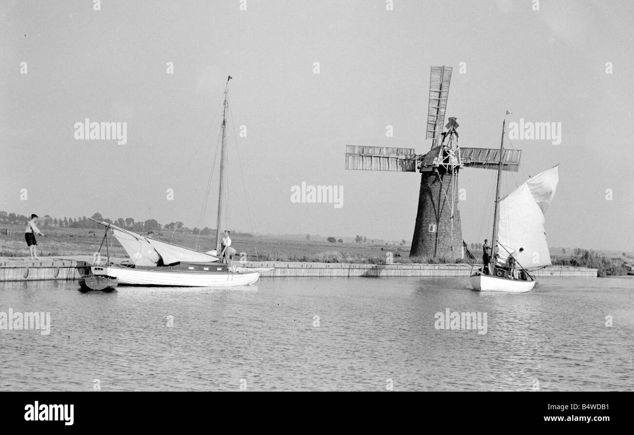 A traditional Norfolk Broad yachts sailing past one of the many windmill water pumps that help with the drainage of the Broads Landscape Sailing Circa 1935 Boats Stock Photo