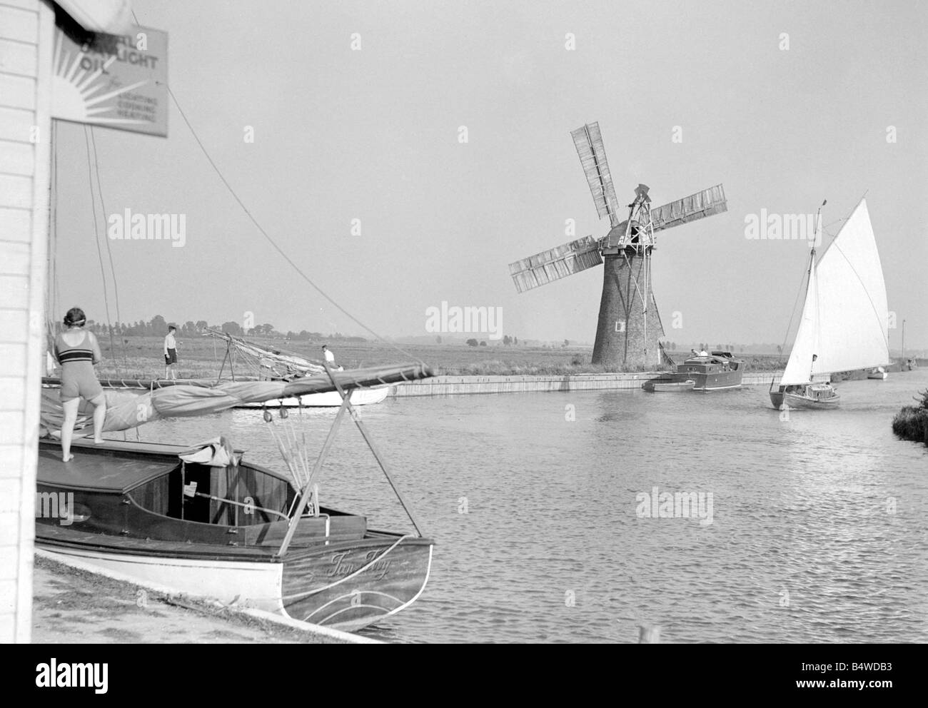 Traditional Norfolk Broad yachts sailing past one of the many windmill water pumps that help with the drainage of the Broads Landscape Sailing Circa 1935 Boats Stock Photo