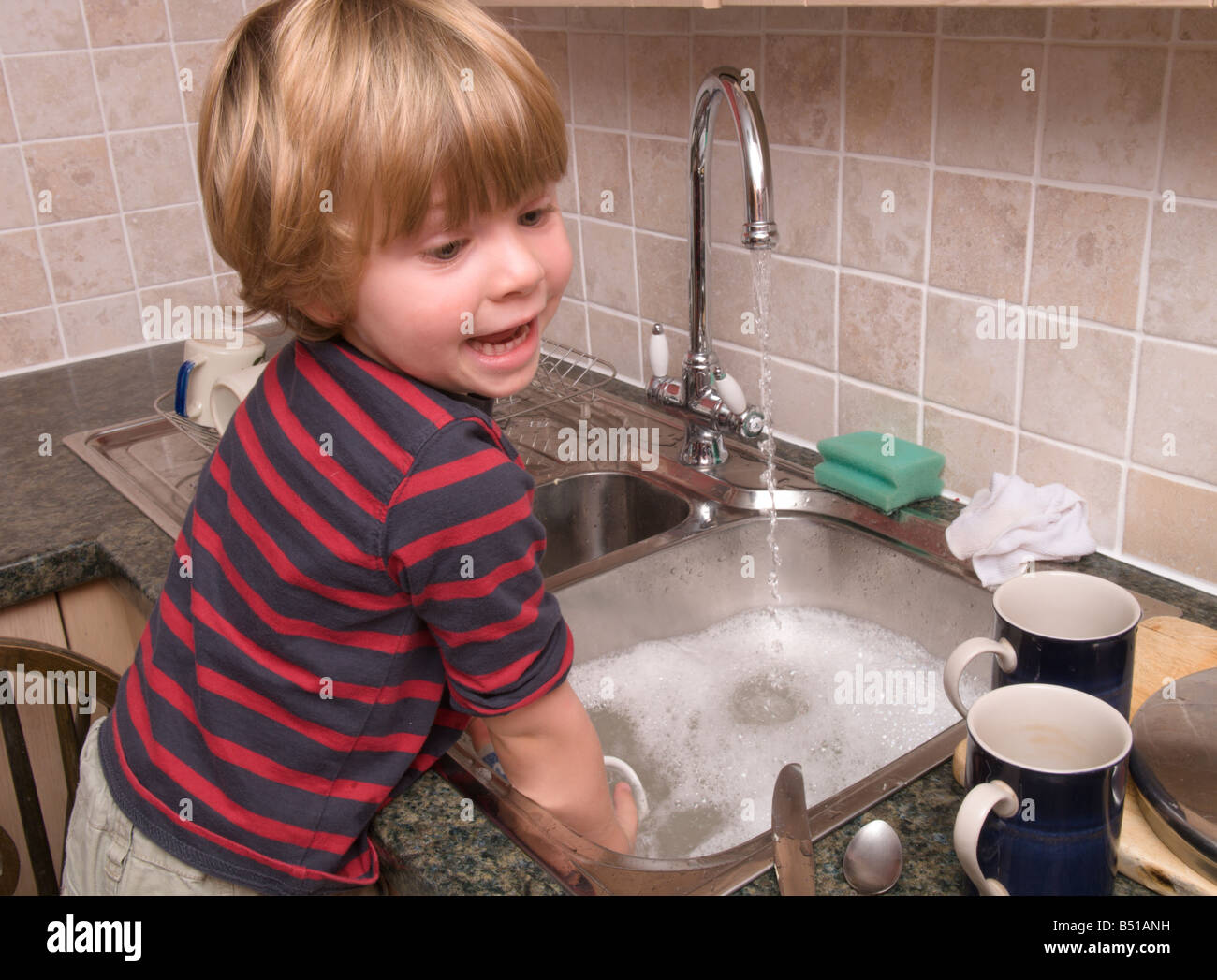young child washing the dishes at the kitchen sink standing on a stool with the hot tap running Stock Photo
